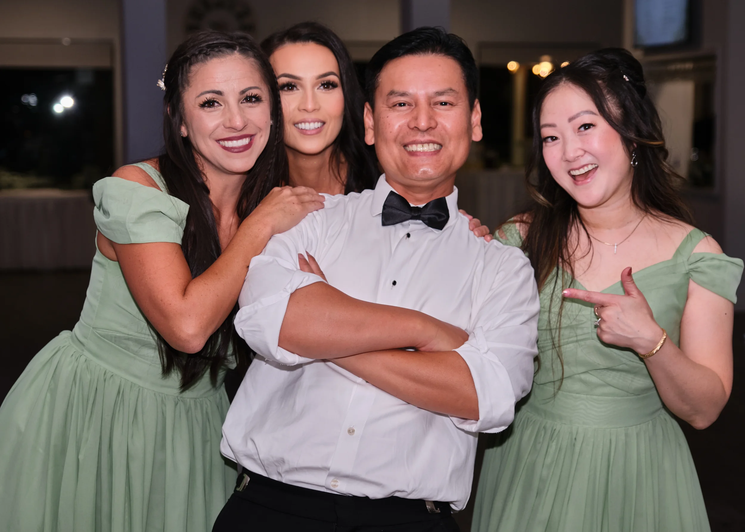 A group of four friends, two women and one man, smiling and posing closely together at a social event. The women are wearing green dresses, and the man is dressed in a white shirt with a black bow tie. They are standing indoors in a warmly lit room.