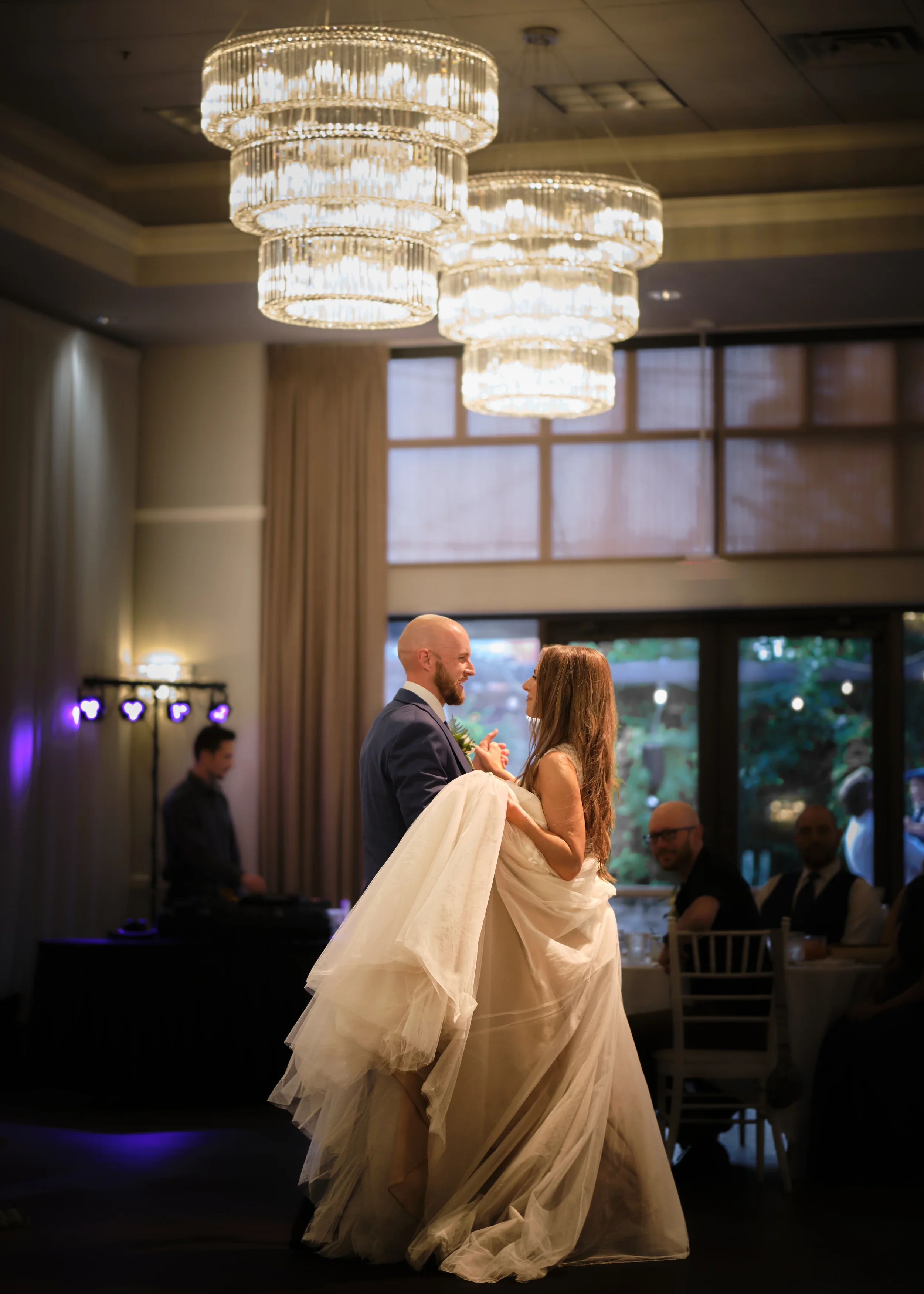 A bride and groom dance together at their wedding reception, holding each other closely in a venue with elegant chandeliers and large windows.