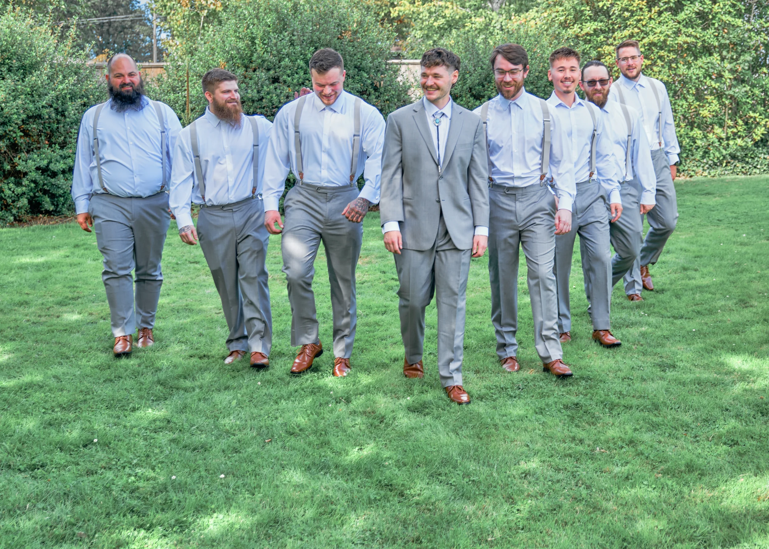 A group of eight men walking in a line outdoors on a grassy area, dressed in formal attire, smiling and enjoying each other's company.