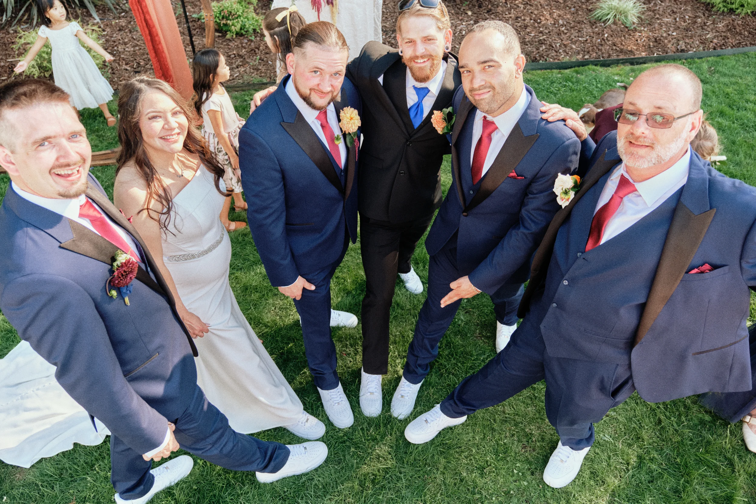 Group of people in suits standing in a circle outdoors, smiling at the camera, with children and a garden in the background.