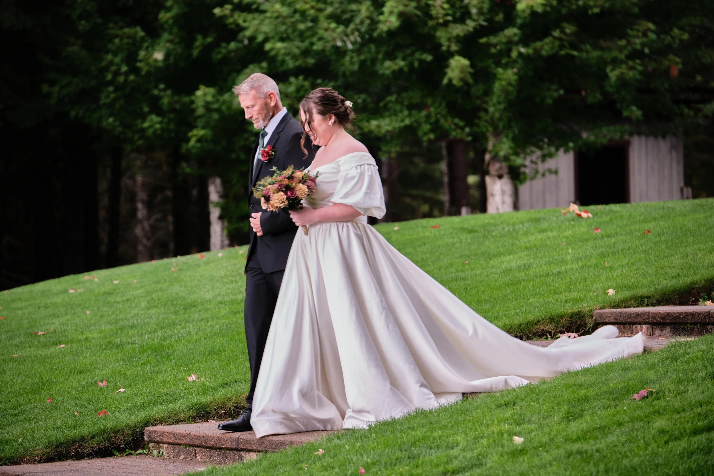 A bride and groom walking on a grassy lawn during a wedding ceremony outdoors, with trees and a small wooden shed in the background.