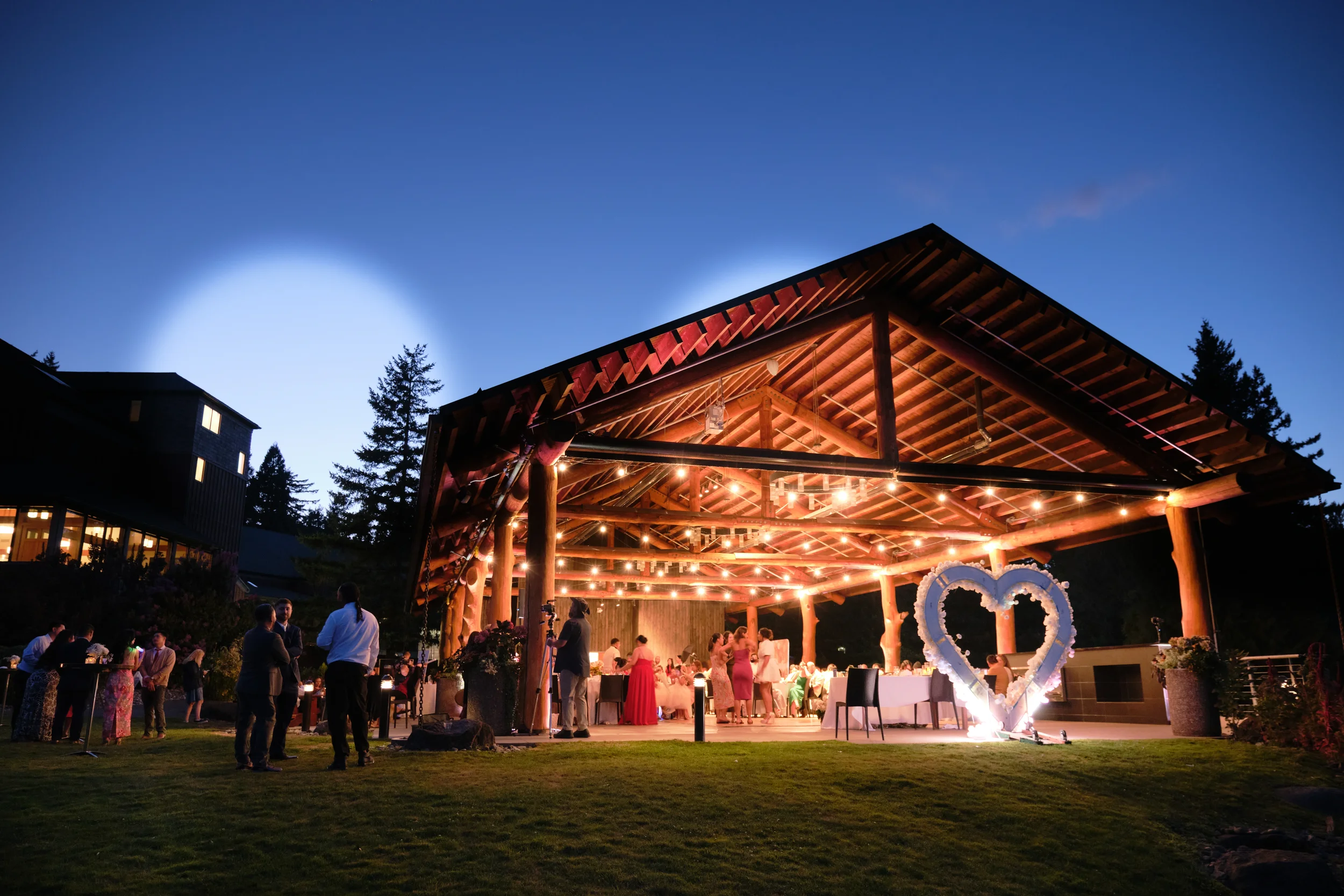 A nighttime outdoor event with people gathered under a wooden pavilion with string lights, celebrating against a dark sky with a bright moon and trees in the background.