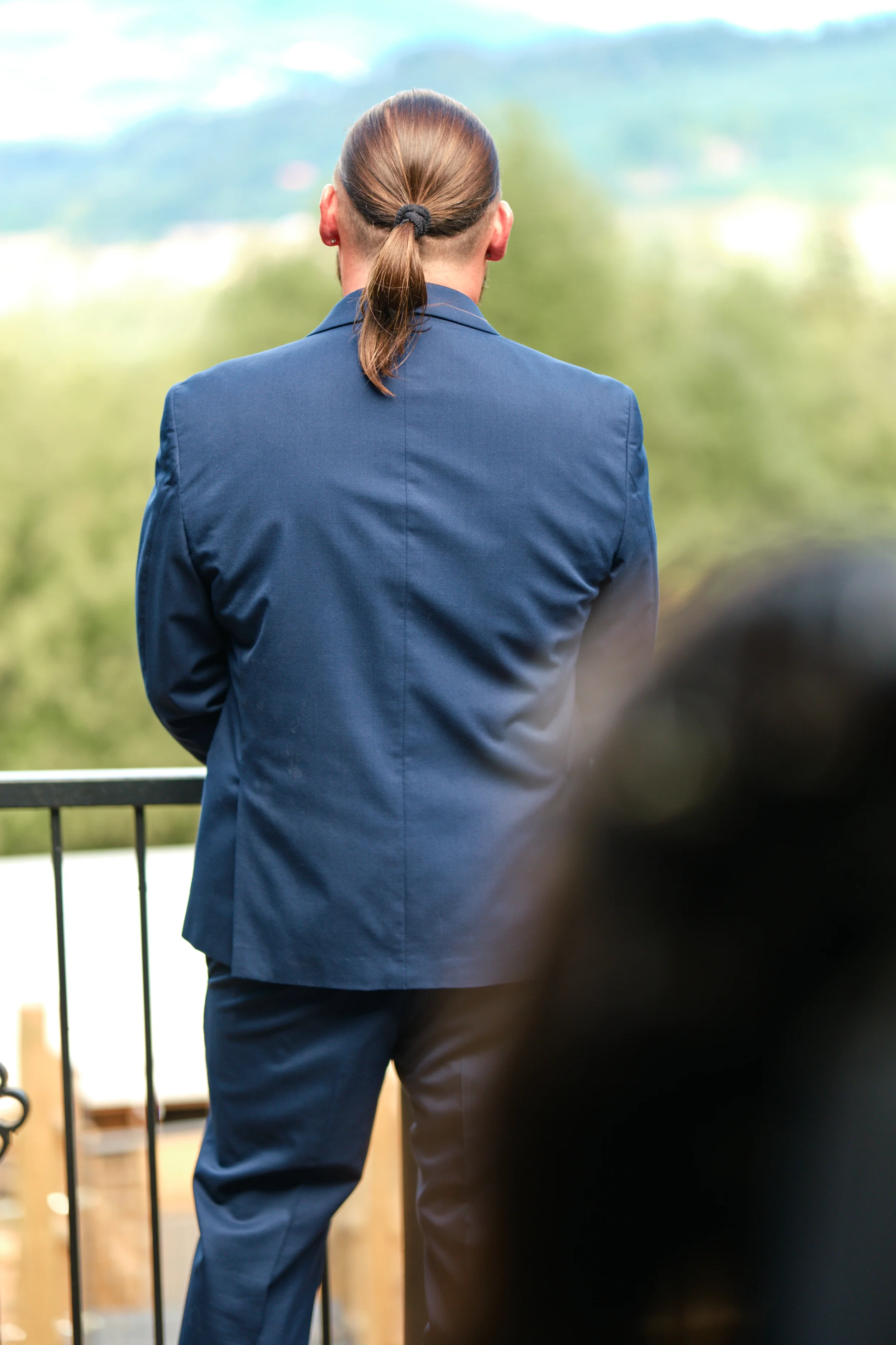 A person with long hair tied back, wearing a blue suit, facing away outdoors with a scenic landscape in the background.