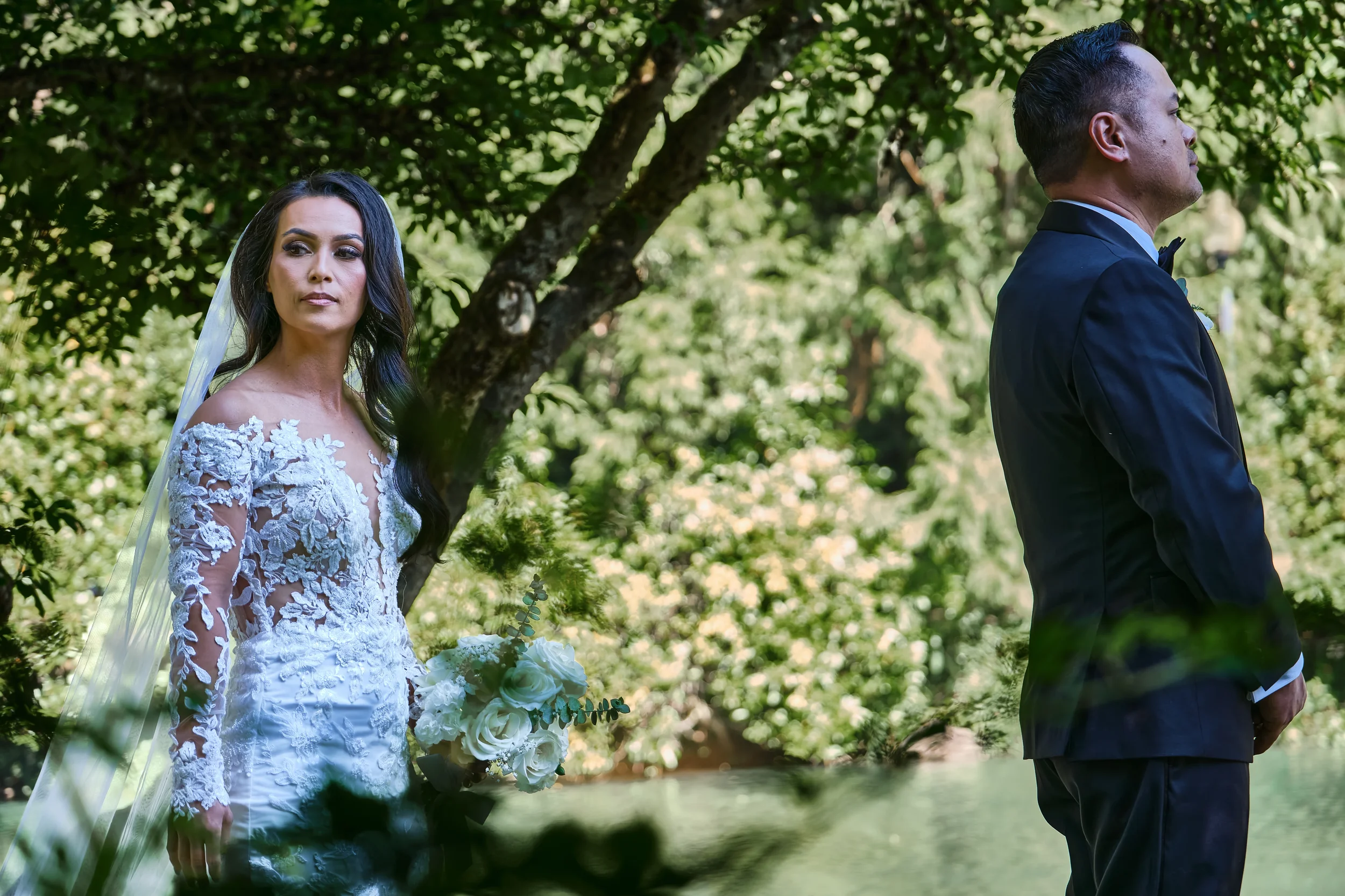 A bride in a white lace wedding gown with long sleeves and a veil holding a bouquet of white roses, standing outdoors next to a groom in a black tuxedo with a bow tie, on a sunny day in a green park with trees.