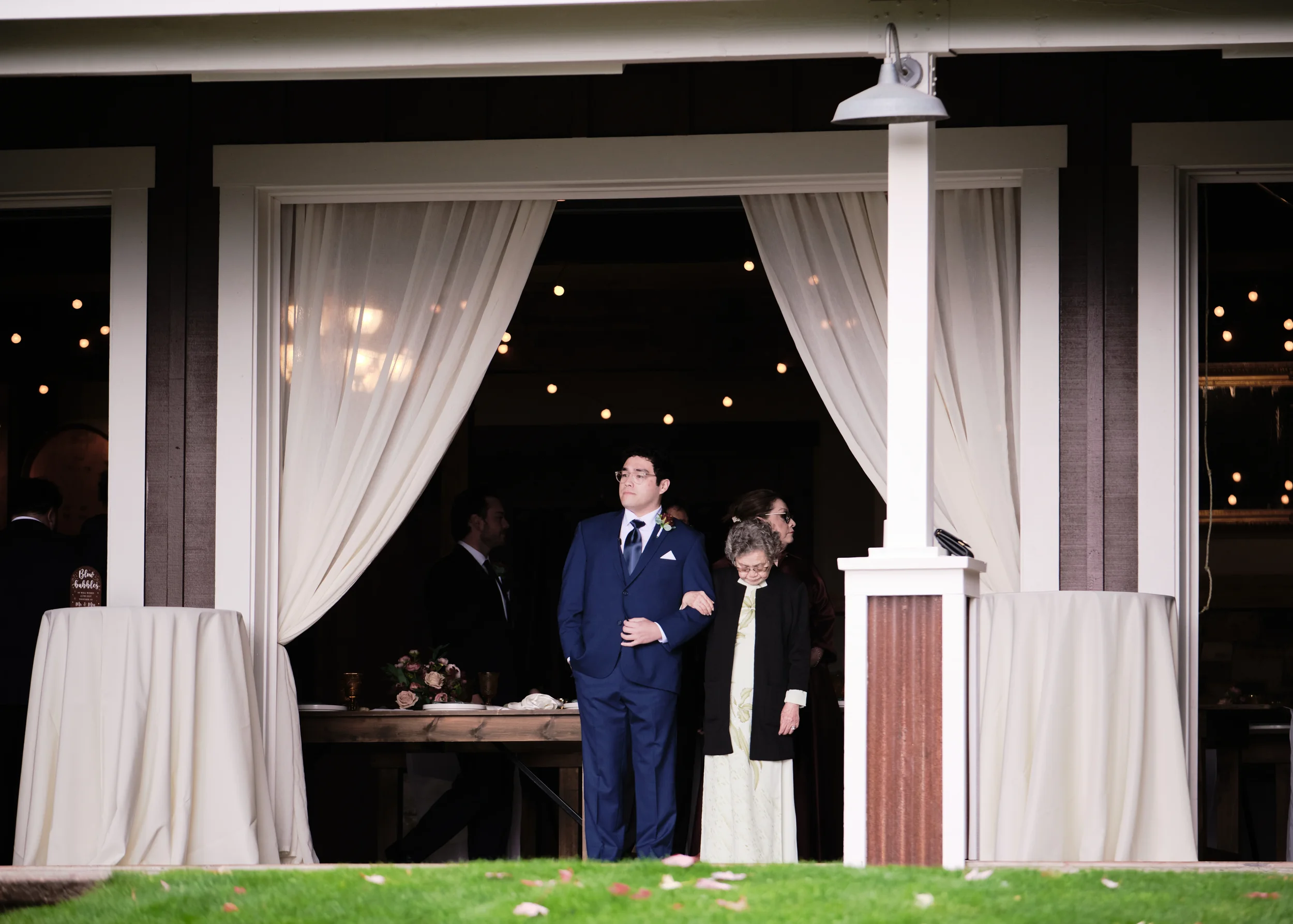 People gather at a wedding reception, with some standing under a covered patio with white curtains, outside a building with large windows and string lights inside, during evening.