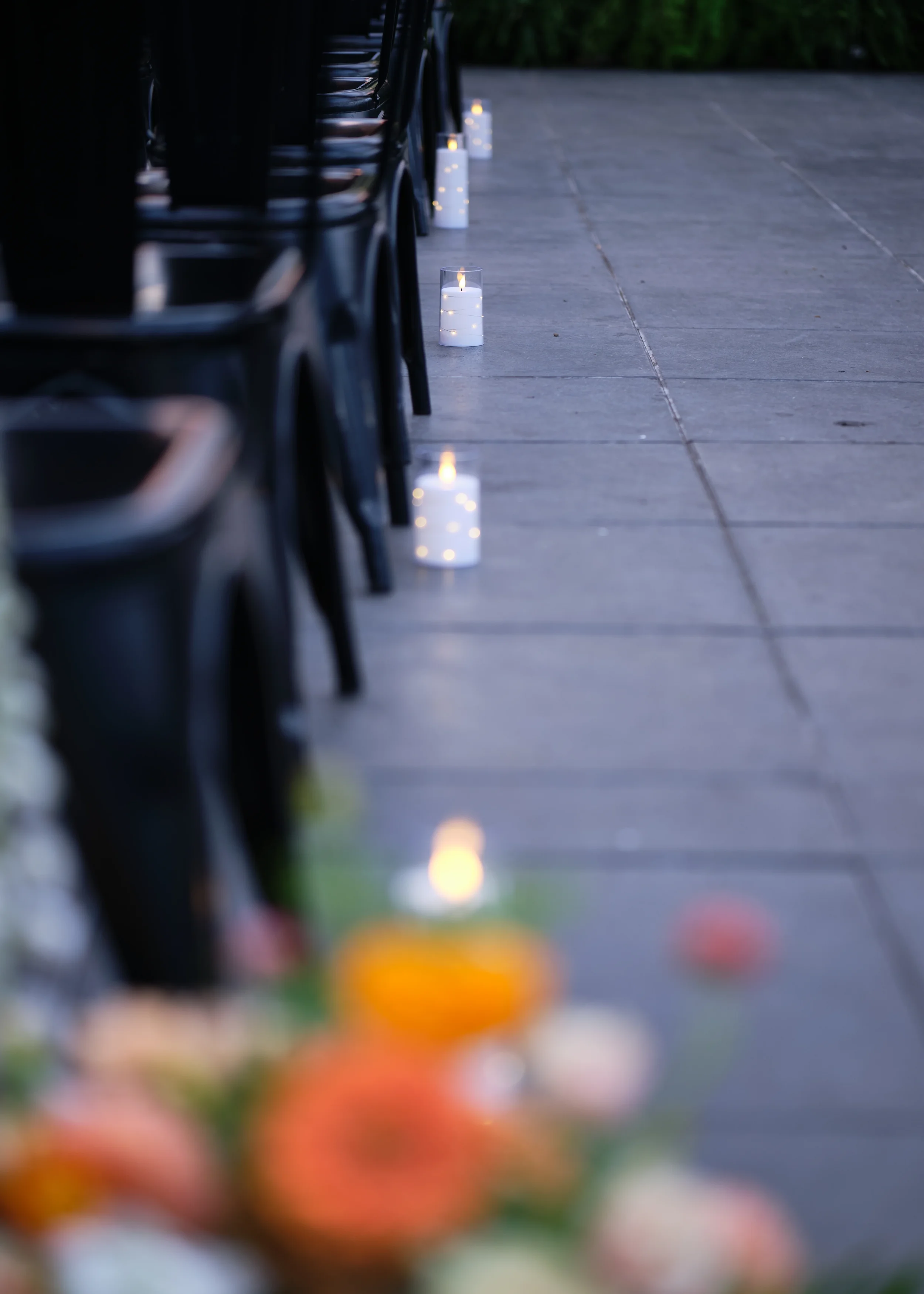 Candles in glass holders placed along a sidewalk at dusk, with black chairs lined up nearby and colorful blurred flowers in the foreground.