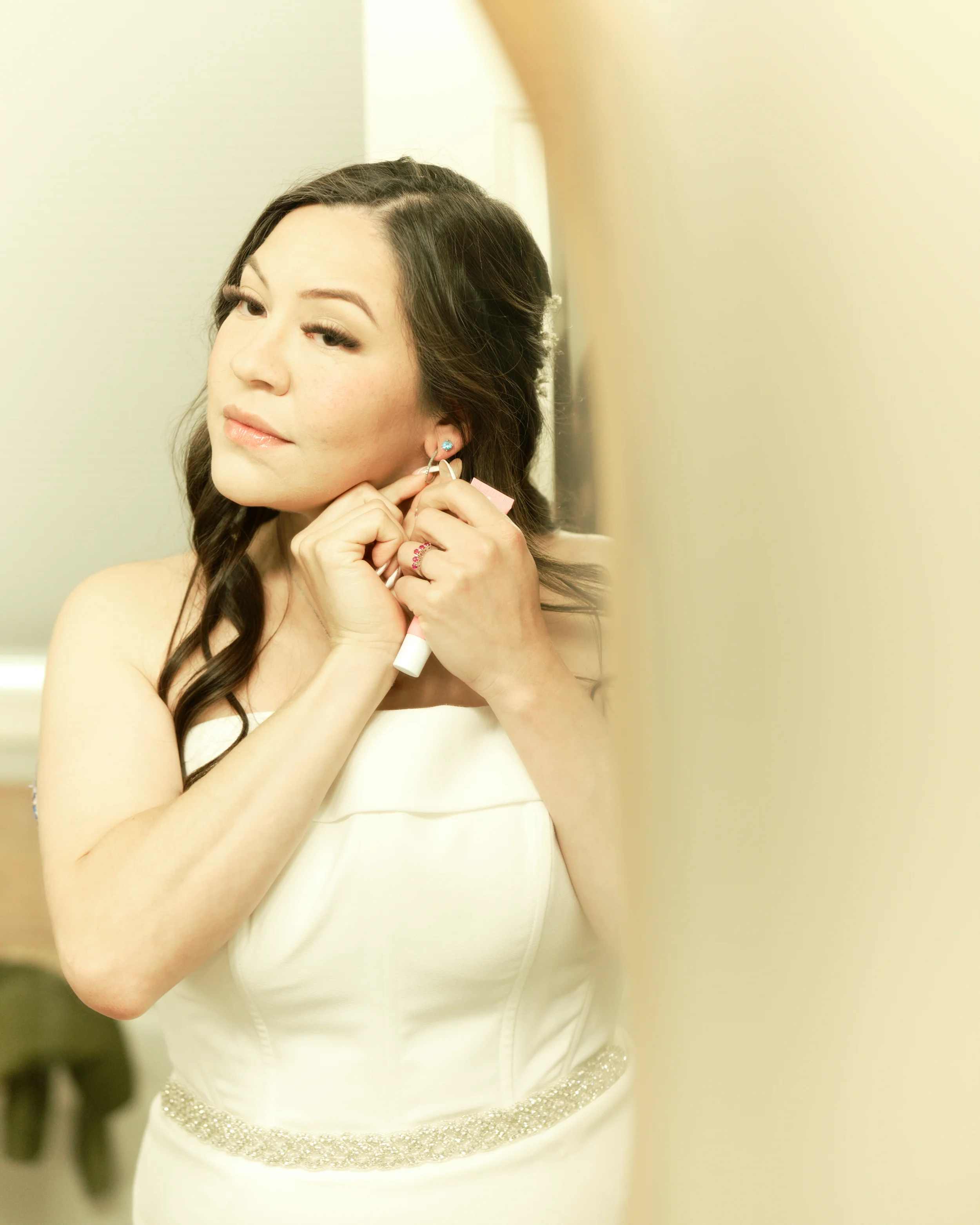 Woman in a white dress putting on an earring in front of a mirror.
