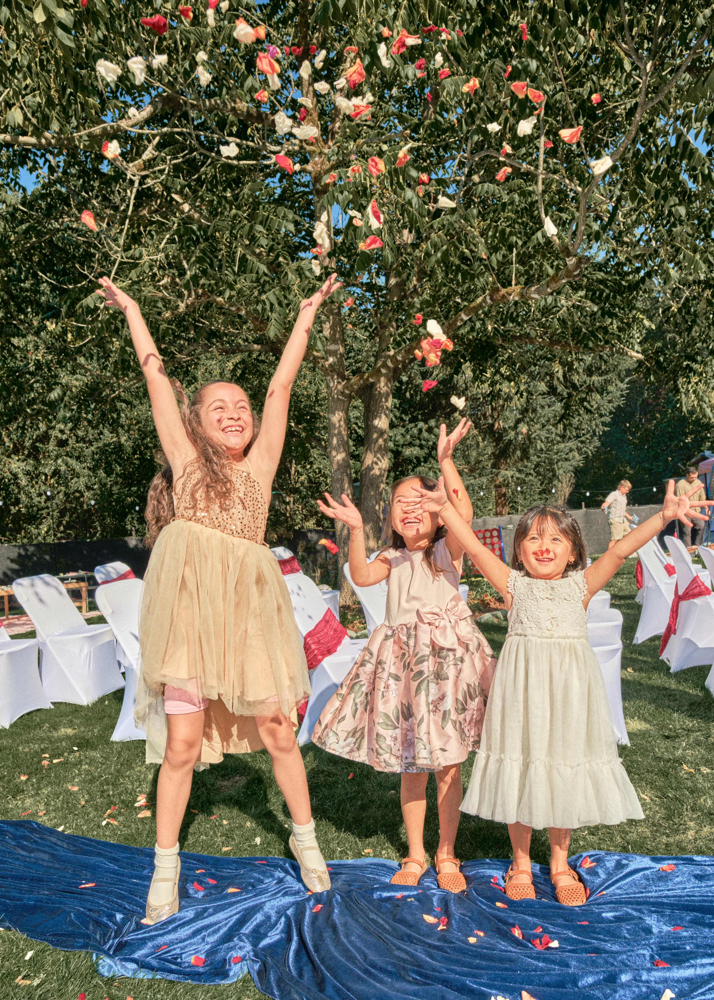 Four young girls standing on a blue tarp outdoors at a celebration, tossing flower petals into the air amid chairs and trees.