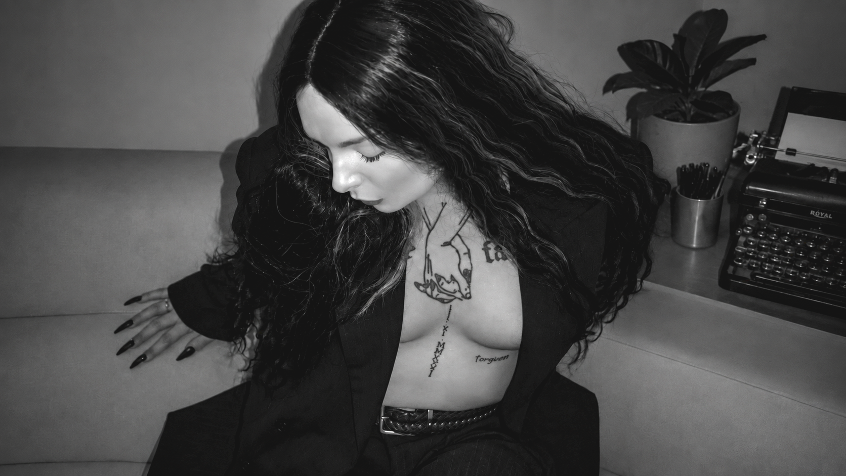 Black and white photograph of a woman with long wavy hair, revealing tattoos on her chest and collarbone area, sitting on a sofa. There is a plant and a vintage typewriter on a table next to her.