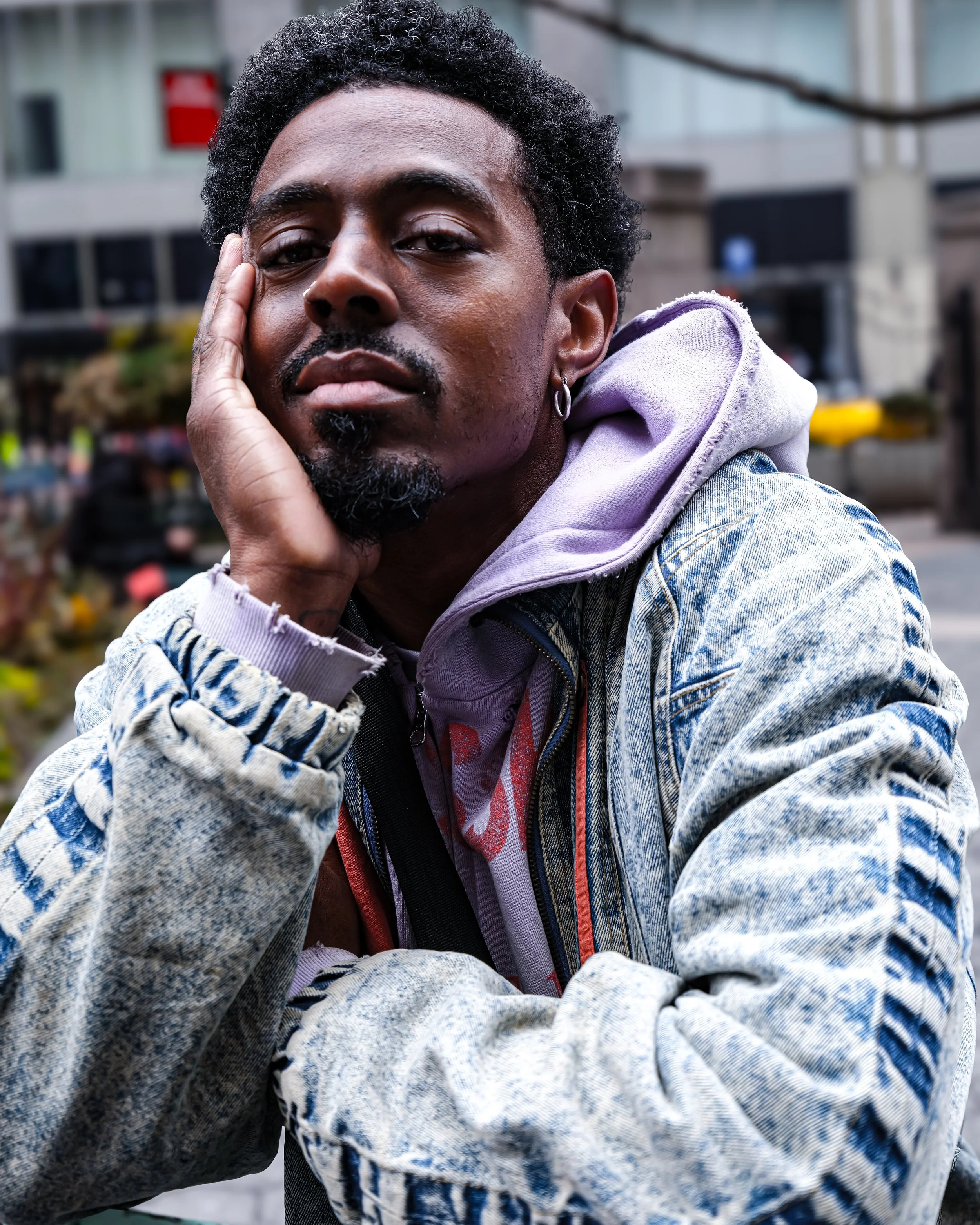 A young Black man with curly hair and a beard, wearing earrings, a purple hoodie, and a denim jacket, resting his face on his hand with a serious expression in an urban outdoor setting.