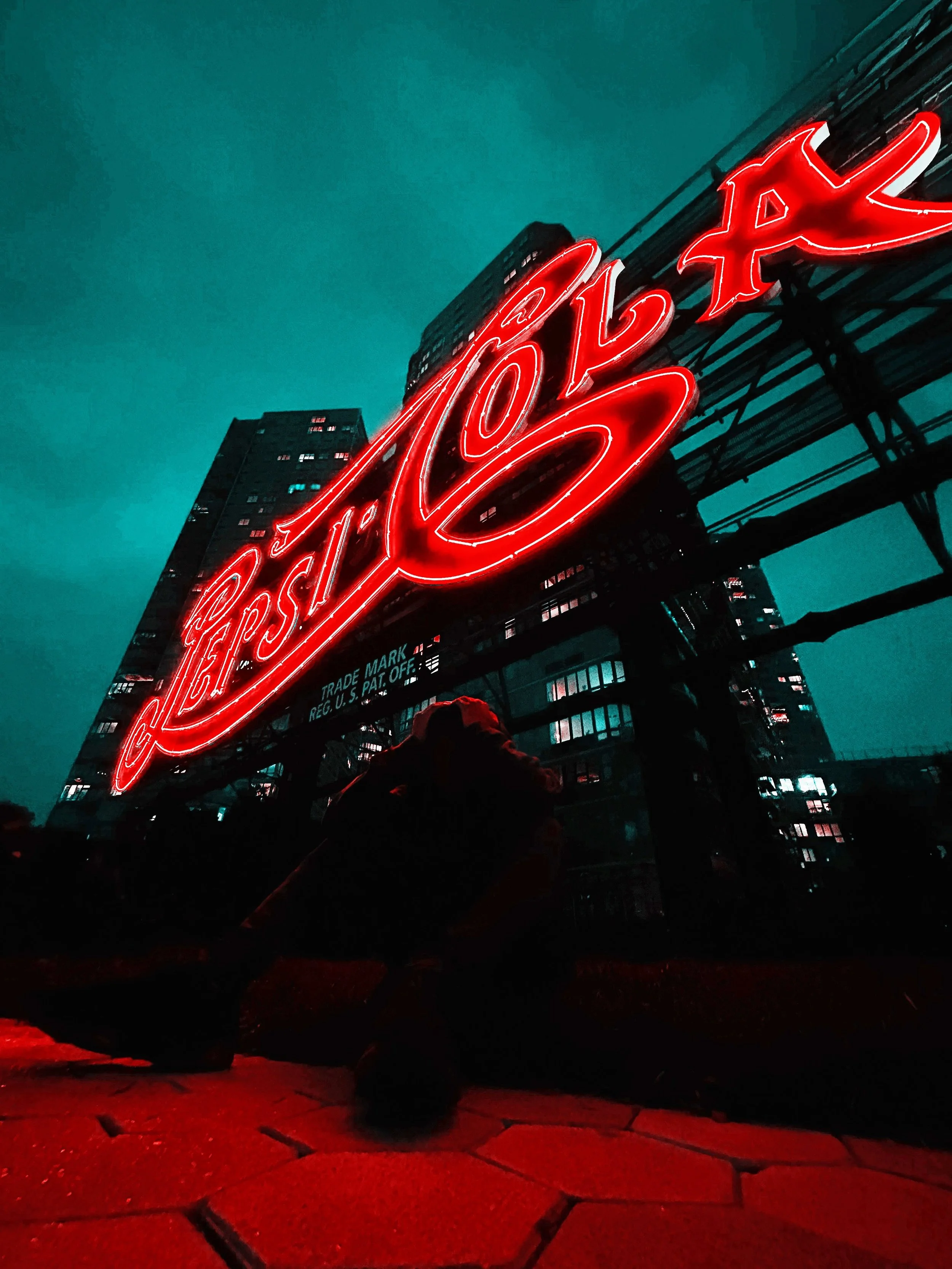 Nighttime city scene with a large illuminated red neon sign on a building that reads 'Pepsi', with the sign's brightness reflecting on red-lit pavement in the foreground.