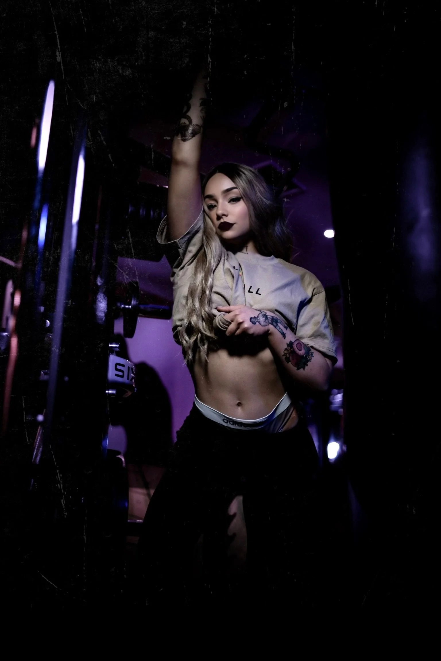 A young woman with tattoos, dark lipstick, long wavy hair, wearing a beige shirt and black shorts, standing in a dimly lit room with purple neon lighting. She is looking at the camera with one arm raised and touching a mirror or dark surface, and the other hand resting on her stomach.