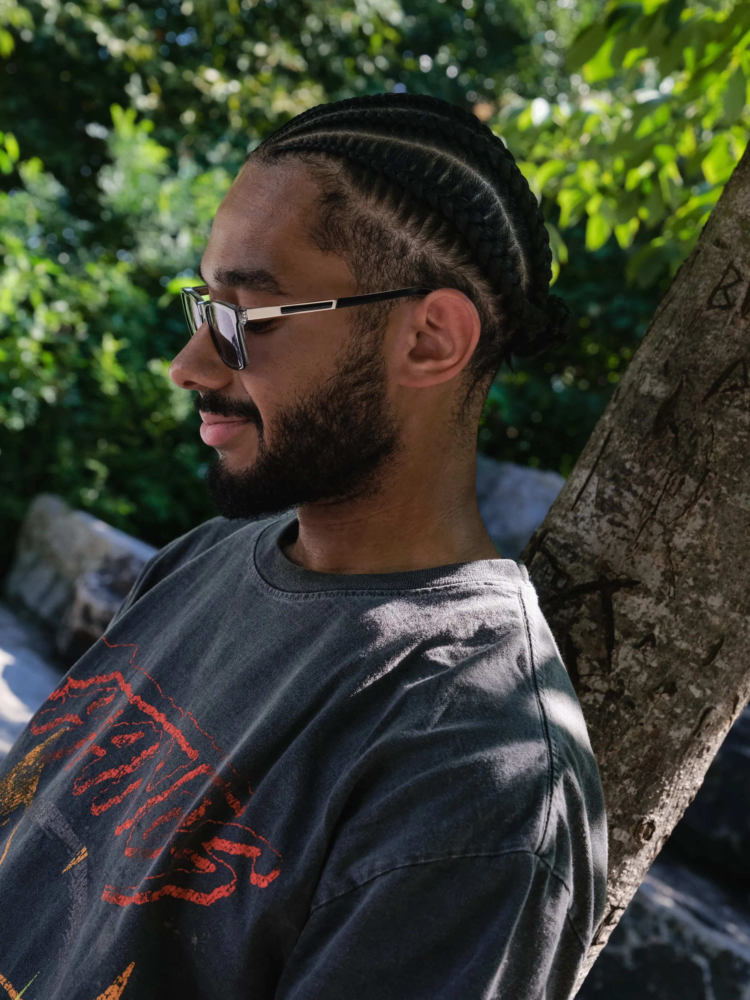 A man with sunglasses and a beard leaning against a tree in a forested area.