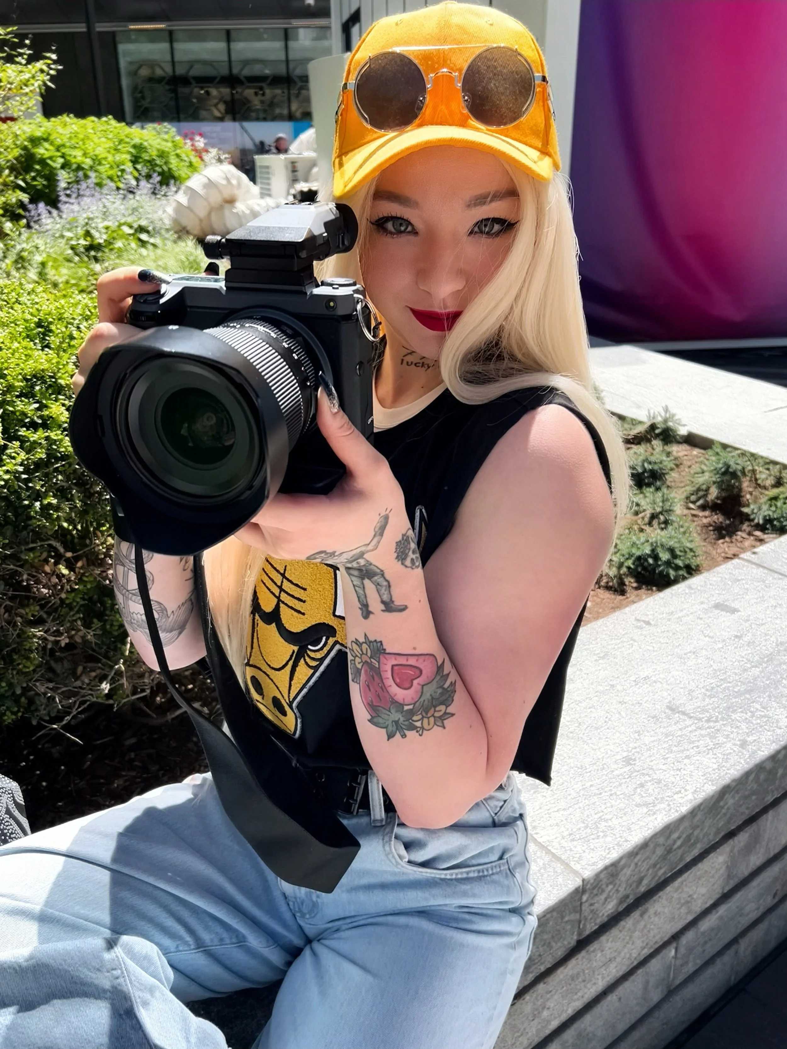 A woman with blonde hair wearing a yellow cap with sunglasses on top, holding a camera while sitting outdoors on a concrete bench with a background of plants and a modern building.