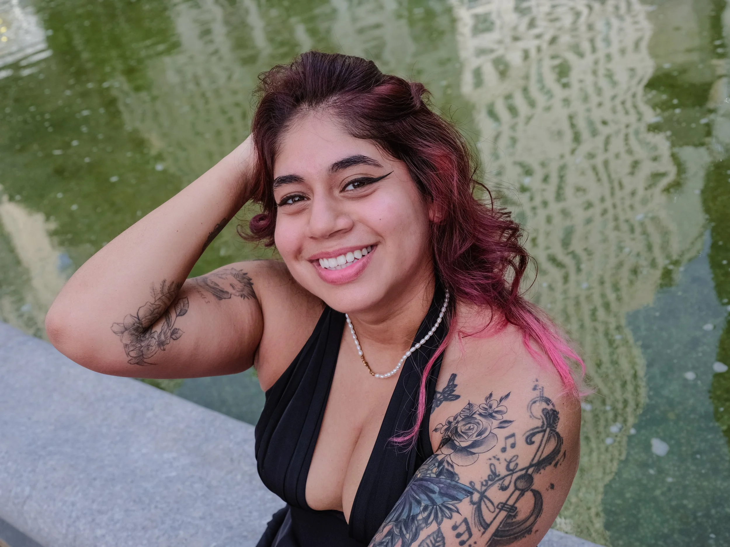 Close-up of a smiling woman with dark and pink hair, tattoos on her arm and shoulder, wearing a black dress and pearl necklace, outdoors near water with green reflections.