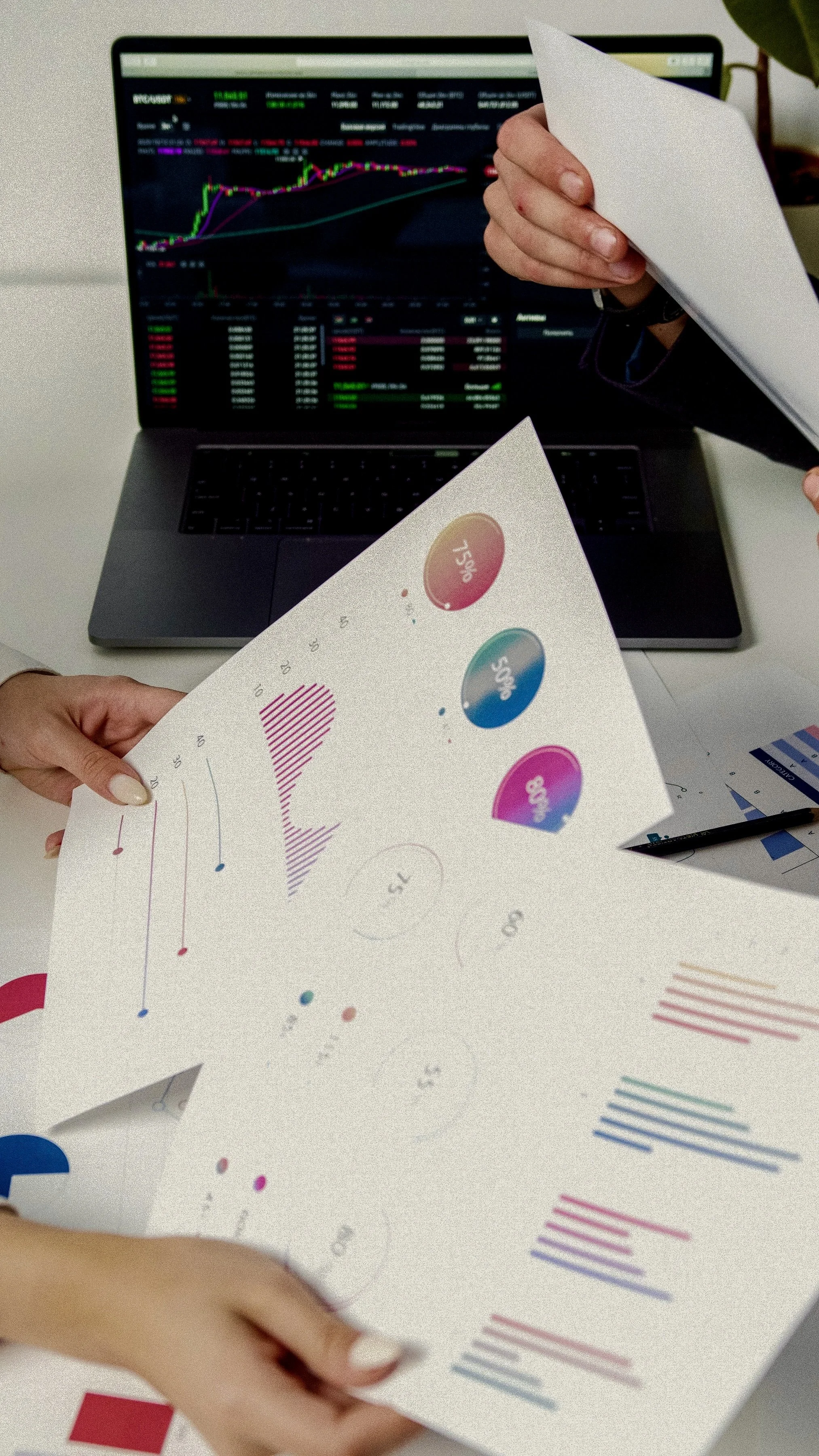 A person is holding printed data charts and graphs with colorful lines and circles, in front of a laptop displaying a stock market or financial trading chart on the screen, with a hand partially visible.