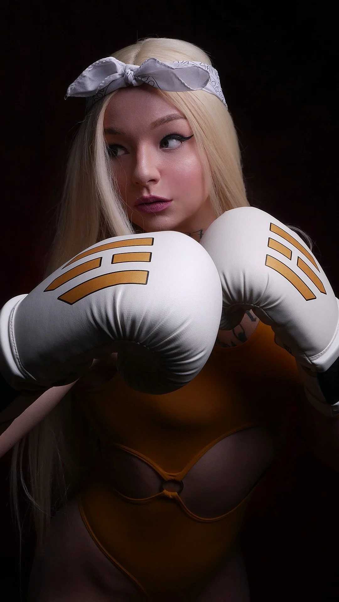 A woman with platinum blonde hair wearing a white bandana, yellow sports bra, and white boxing gloves with gold accents, striking a boxing pose against a dark background.