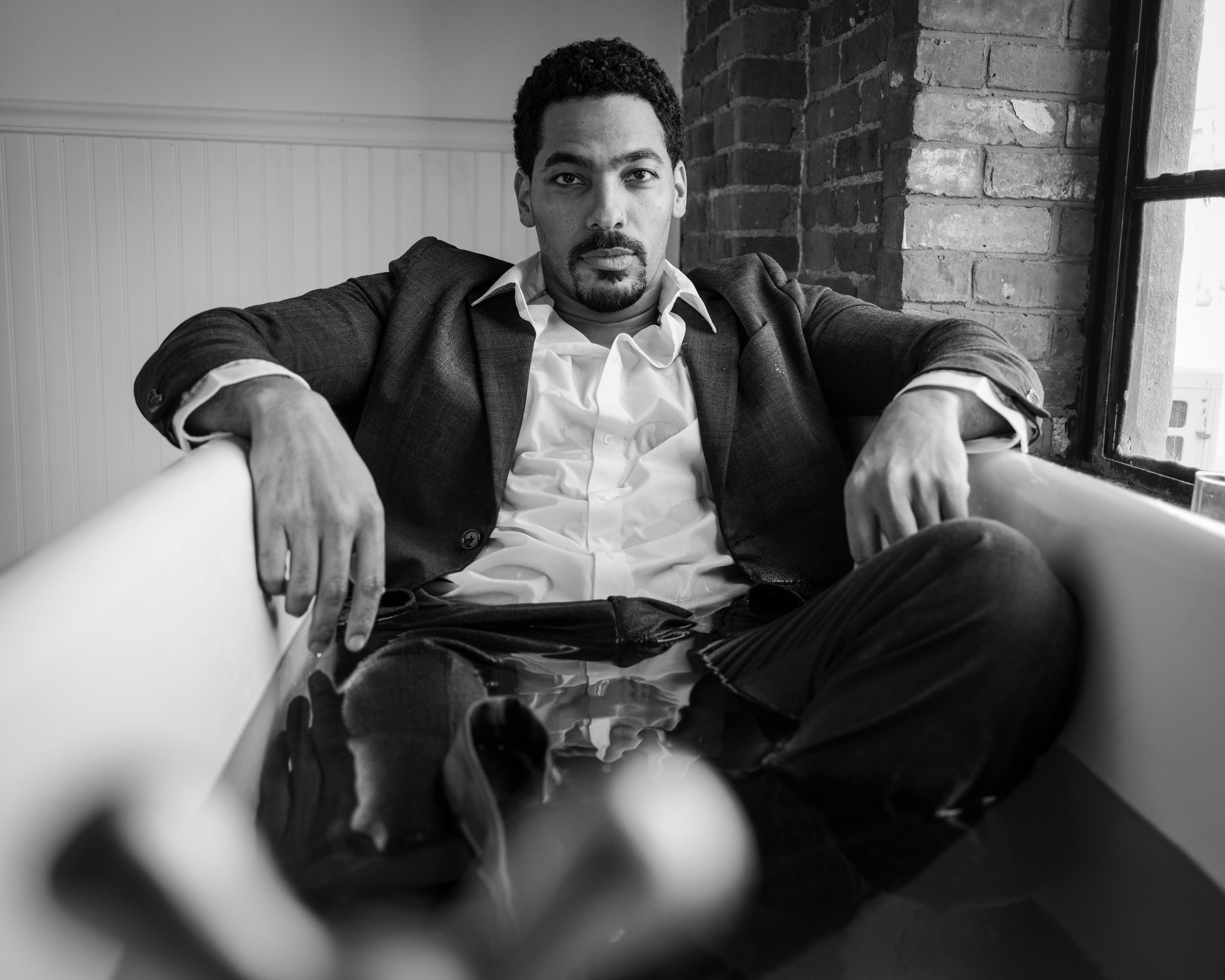 Black and white photo of a man with curly hair and a mustache sitting indoors near a brick wall and a window, wearing a suit jacket over a shirt, looking serious.