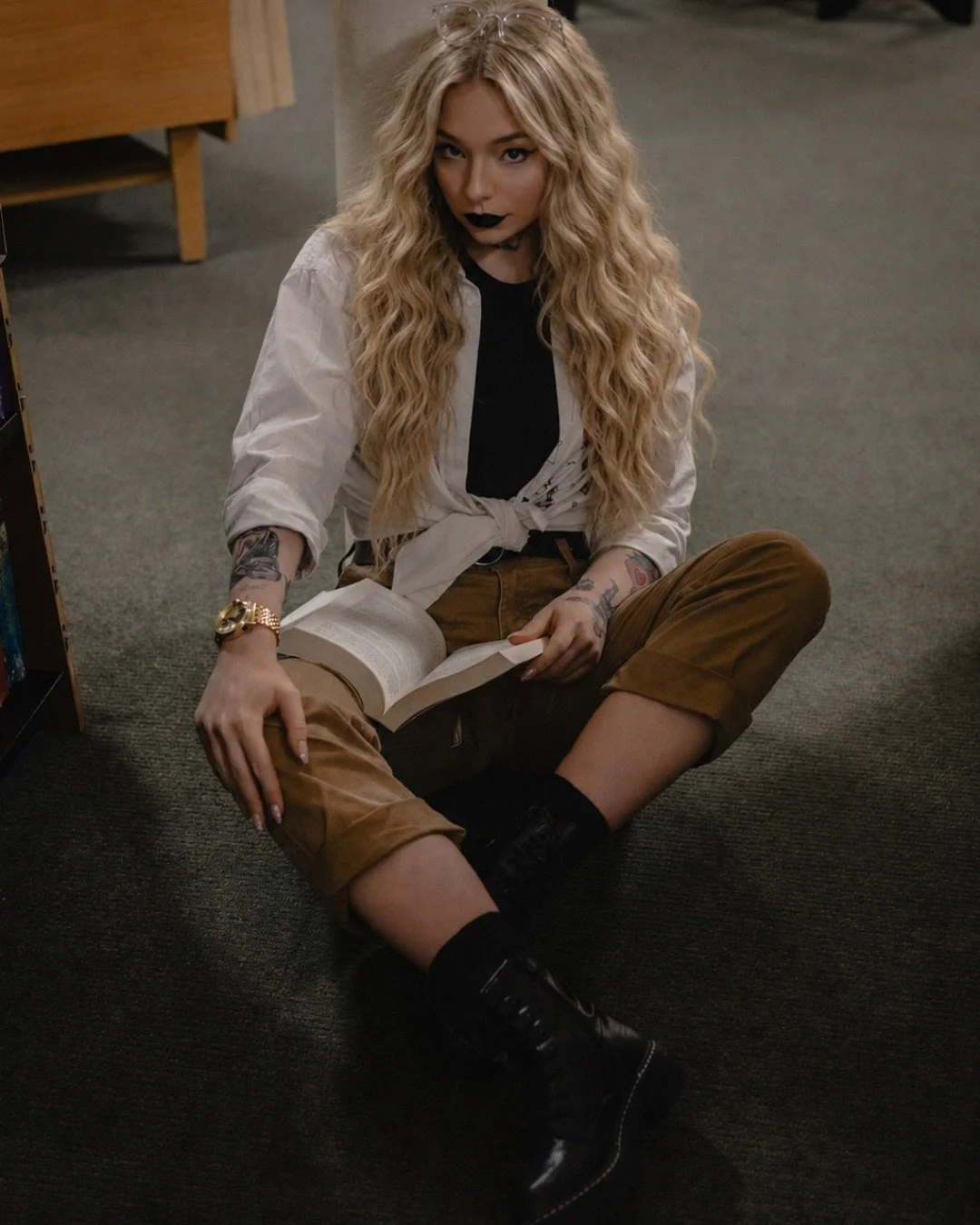 A woman with long, curly blonde hair sitting on the floor of a library, reading a book, wearing a white tied-up shirt, black top, brown pants, black boots, and dark lipstick.
