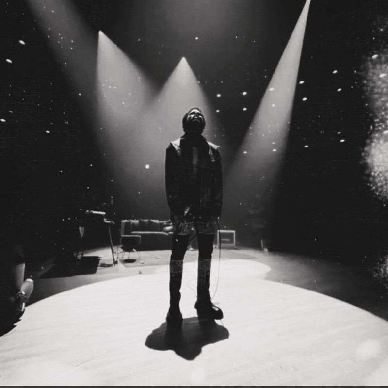A person standing alone on a stage illuminated by three spotlights, with a cosmic background and musical equipment nearby.