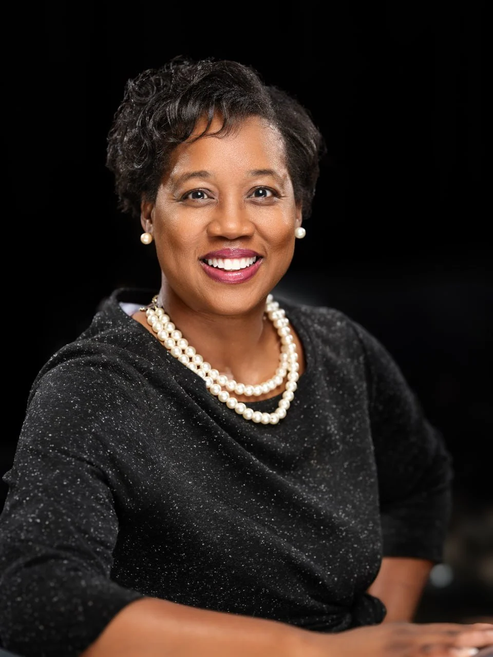 Portrait of a smiling African American woman wearing pearl earrings, a pearl necklace, and a black dress with subtle sparkle, against a black background.