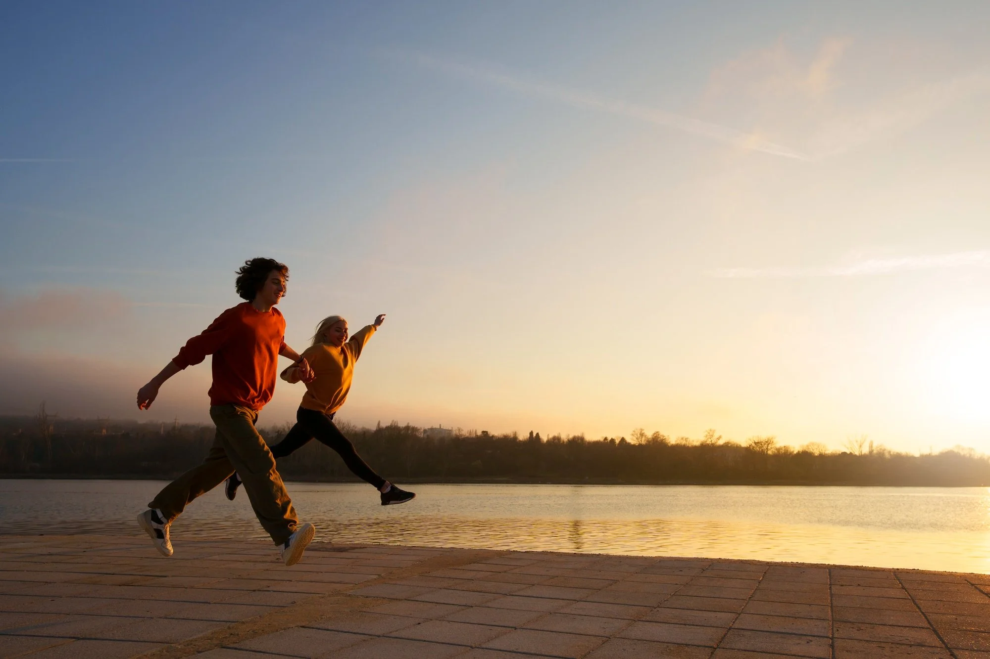 Two people running along a lakeside at sunset, with a clear sky and some clouds in the background.