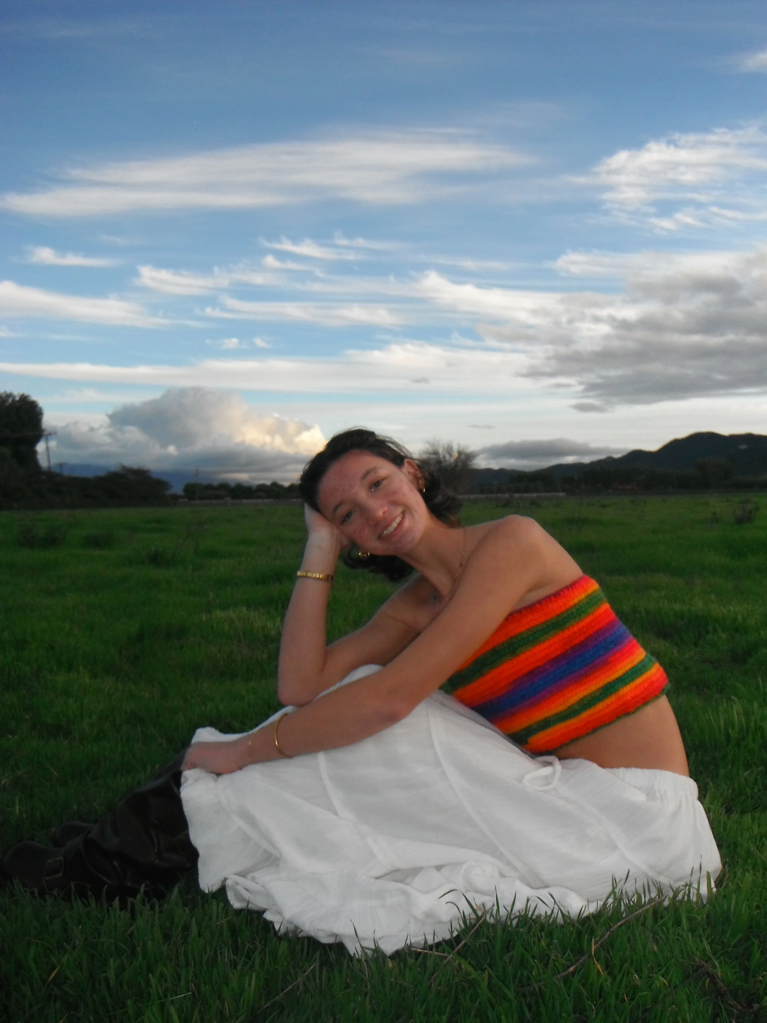 A woman with short dark hair, smiling, sitting on a grassy field during a cloudy day, wearing a colorful striped crop top and a white long skirt.