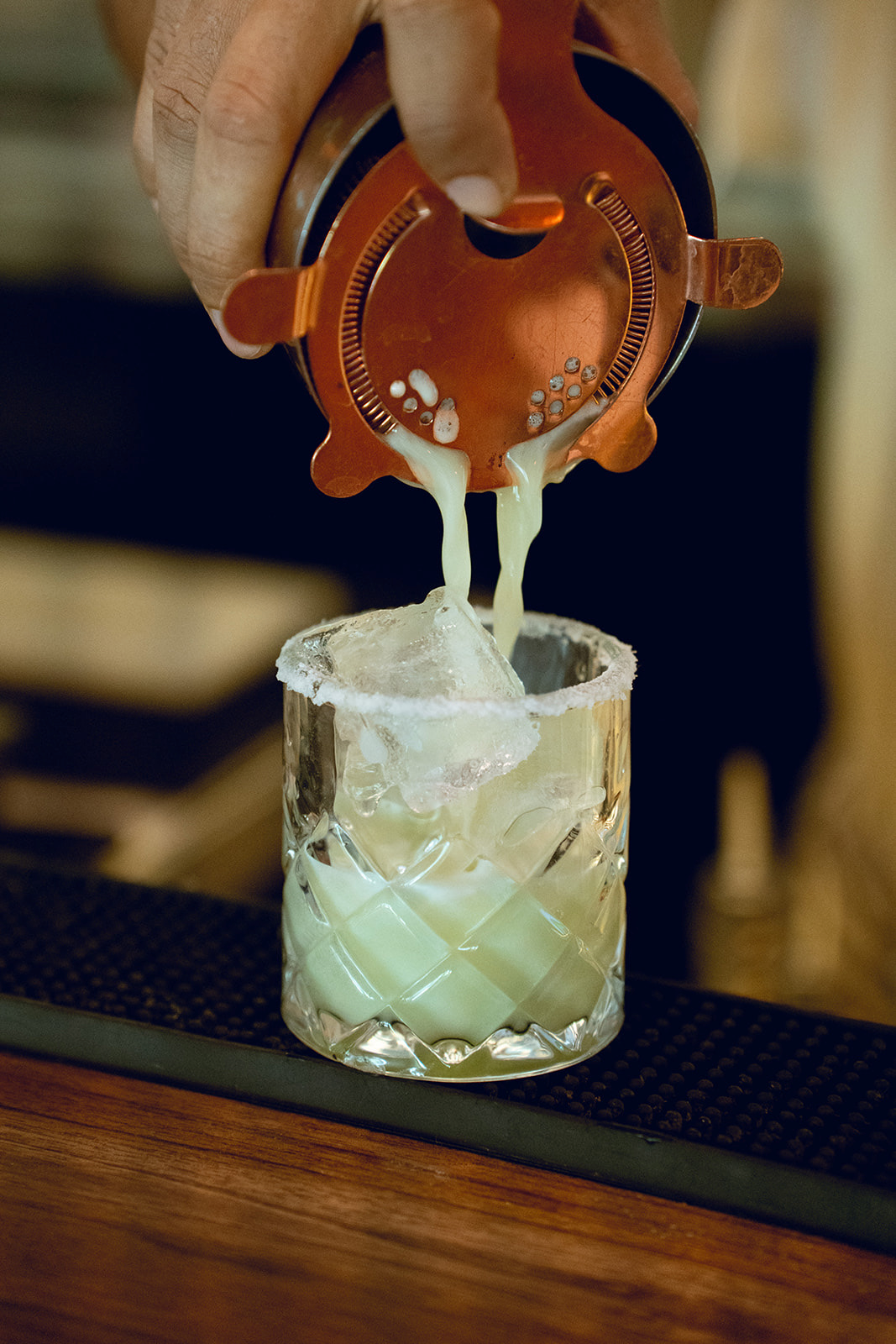 A bartender's hand pouring a creamy white drink from a copper cocktail shaker into a glass with ice, with the rimming of the glass coated with salt.
