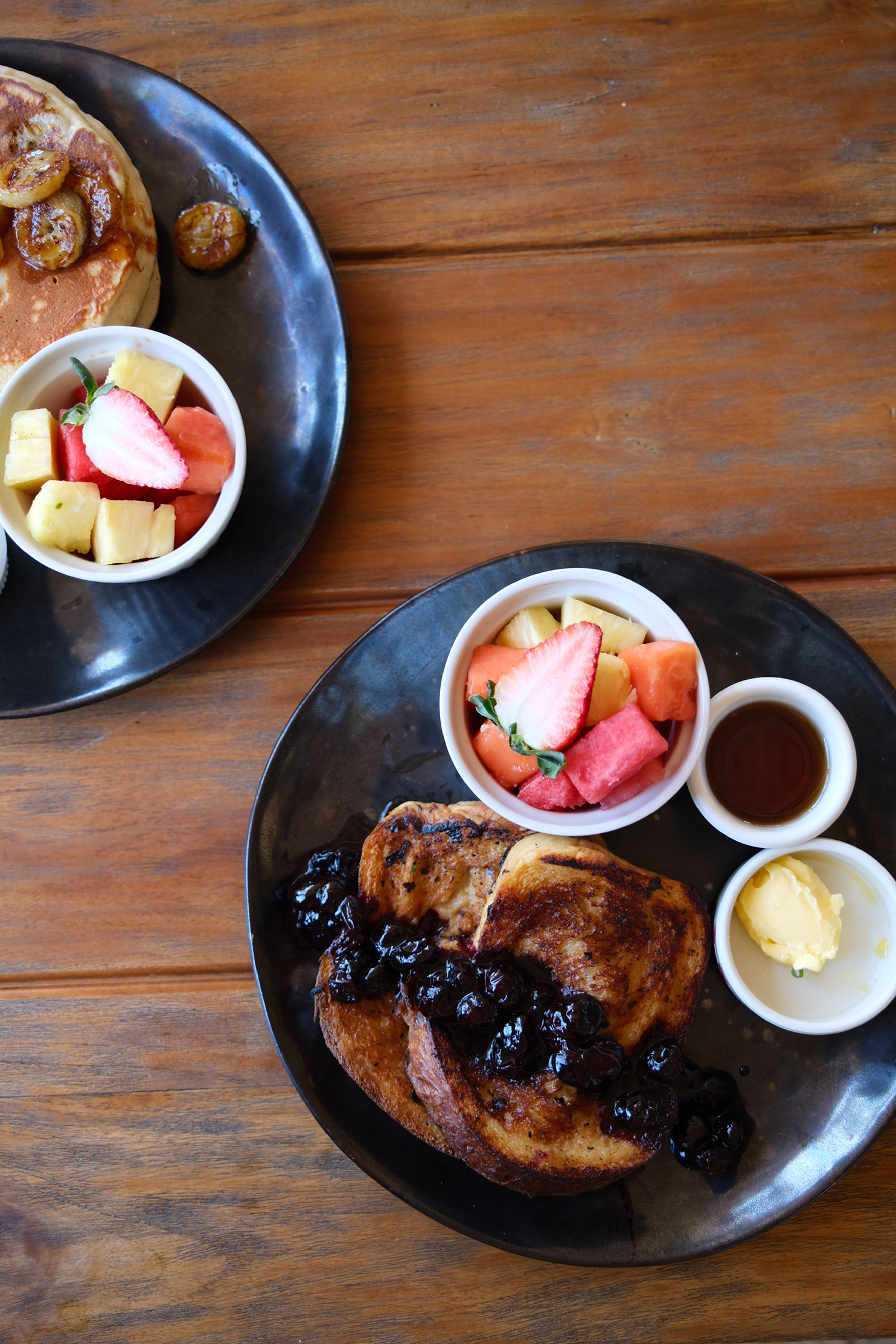 Close-up of a breakfast plate with French toast topped with blueberries and syrup, a small bowl of mixed fresh fruit including strawberries, pineapple, watermelon, and cantaloupe, and small cups of syrup, butter, and butter with herbs, on a wooden table.