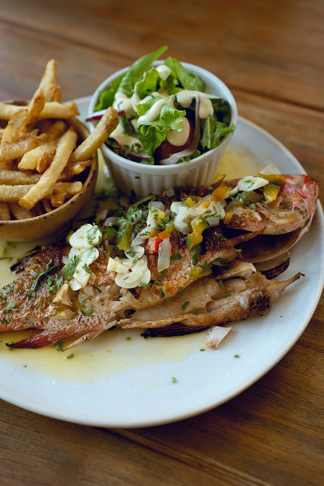 Fried fish topped with chopped vegetables and green onions, served with French fries and a side salad in a small cup on a white plate.