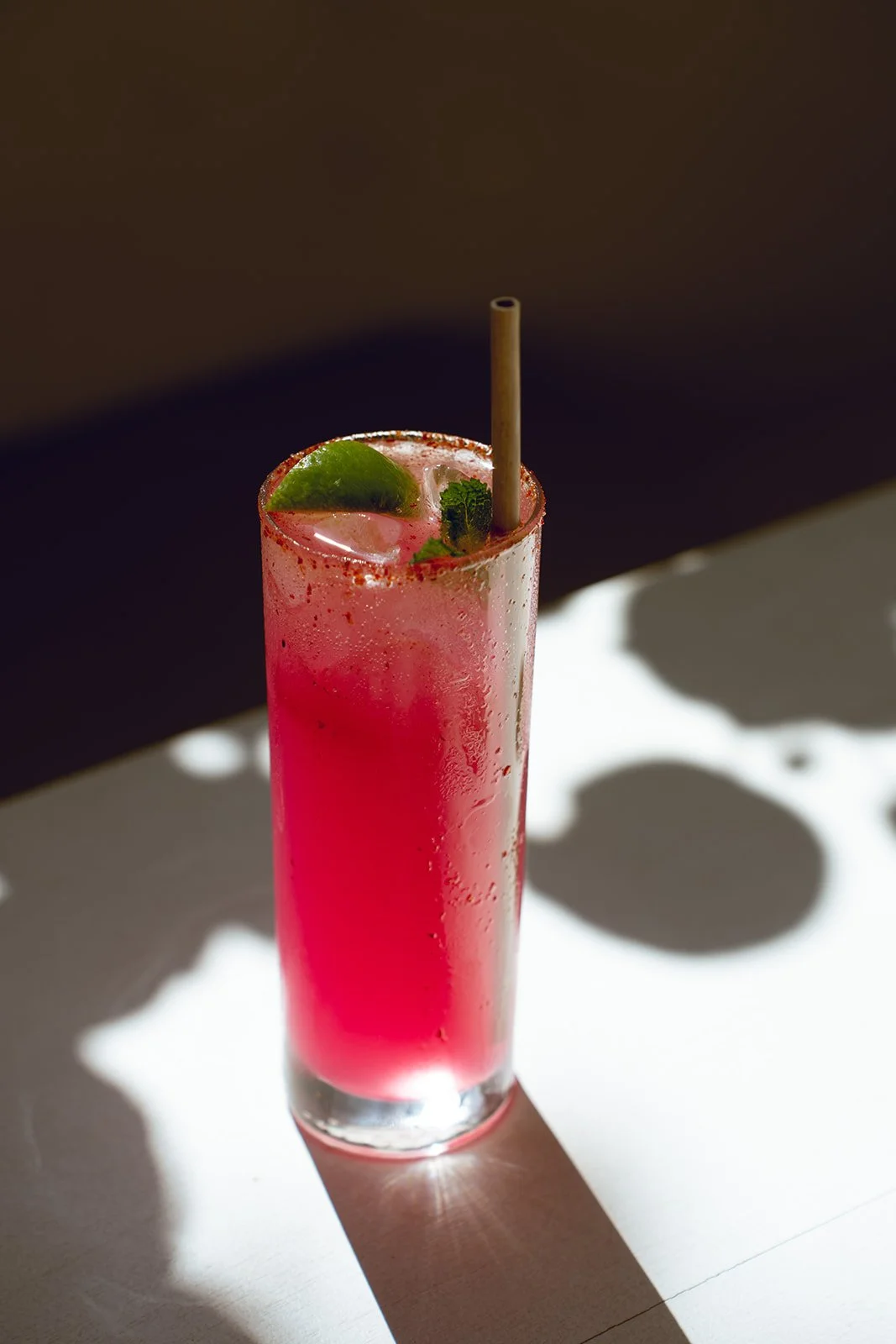 A tall glass of pink cocktail garnished with a lime wedge and mint leaves, with condensation on the glass, placed on a white surface.
