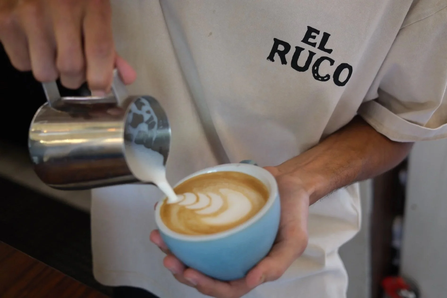 A barista pouring steamed milk into a cup of espresso to make latte art at a coffee shop. The person is wearing a beige shirt with the text 'EL RUC' printed on it.