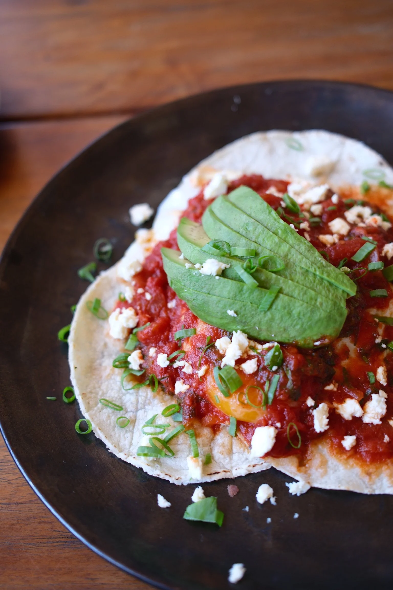 Close-up of a Mexican taco with lettuce, cheese, tomato sauce, and chopped green onions on a black plate.
