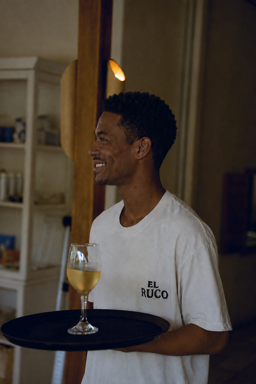 A smiling man wearing a white T-shirt with 'EL RUCO' written on it, holding a black tray with a glass of white wine.