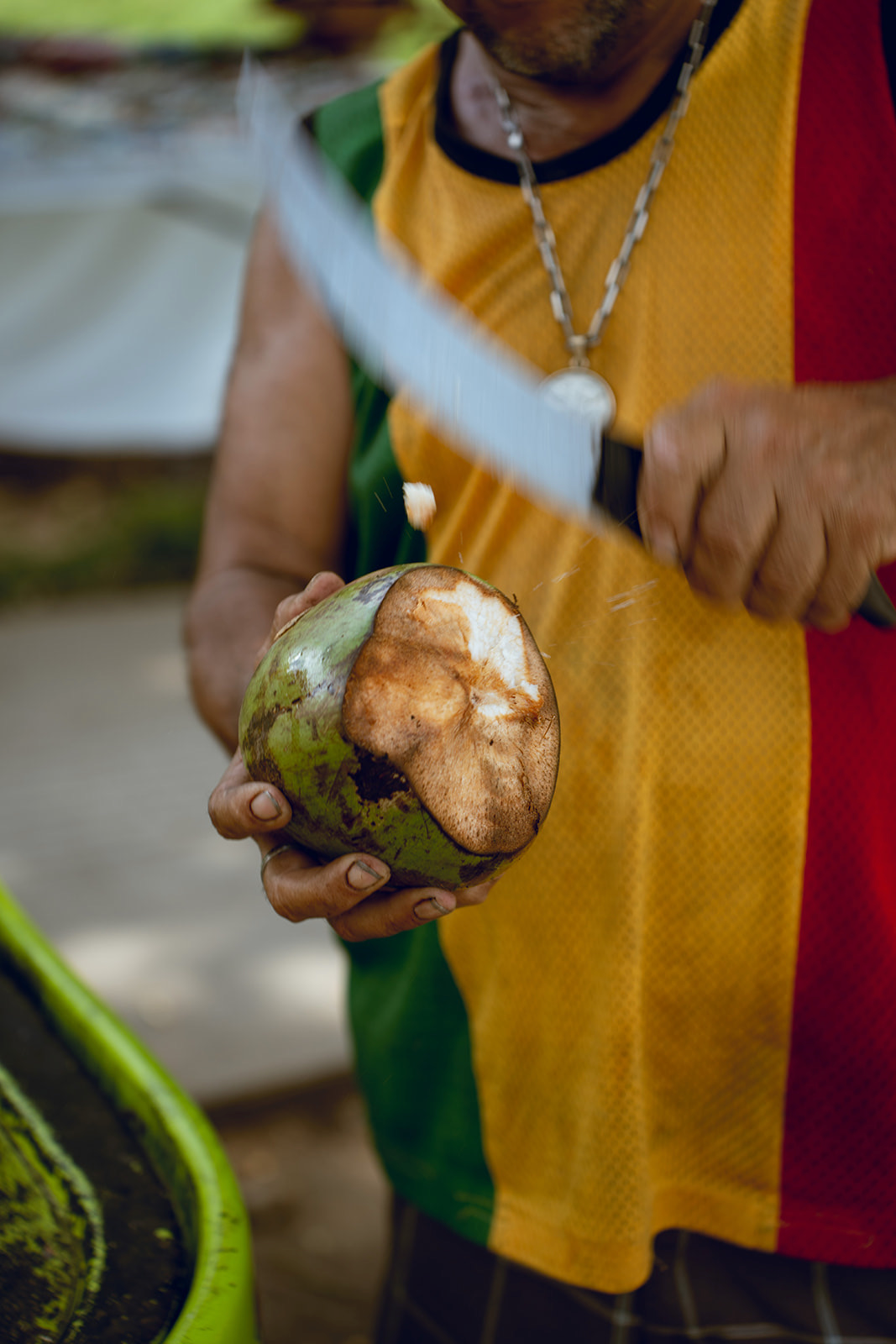 A person holding a green coconut with a brown, partially eaten top, and a hand using a large knife to cut into it.
