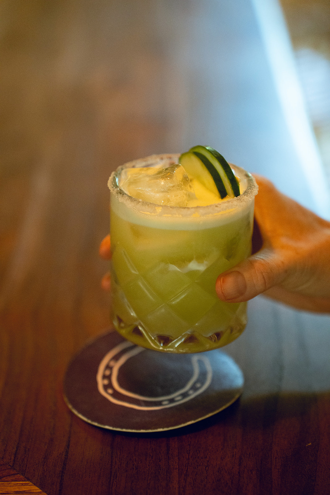 A hand holding a glass of green cocktail with ice and cucumber slices at a bar or restaurant.