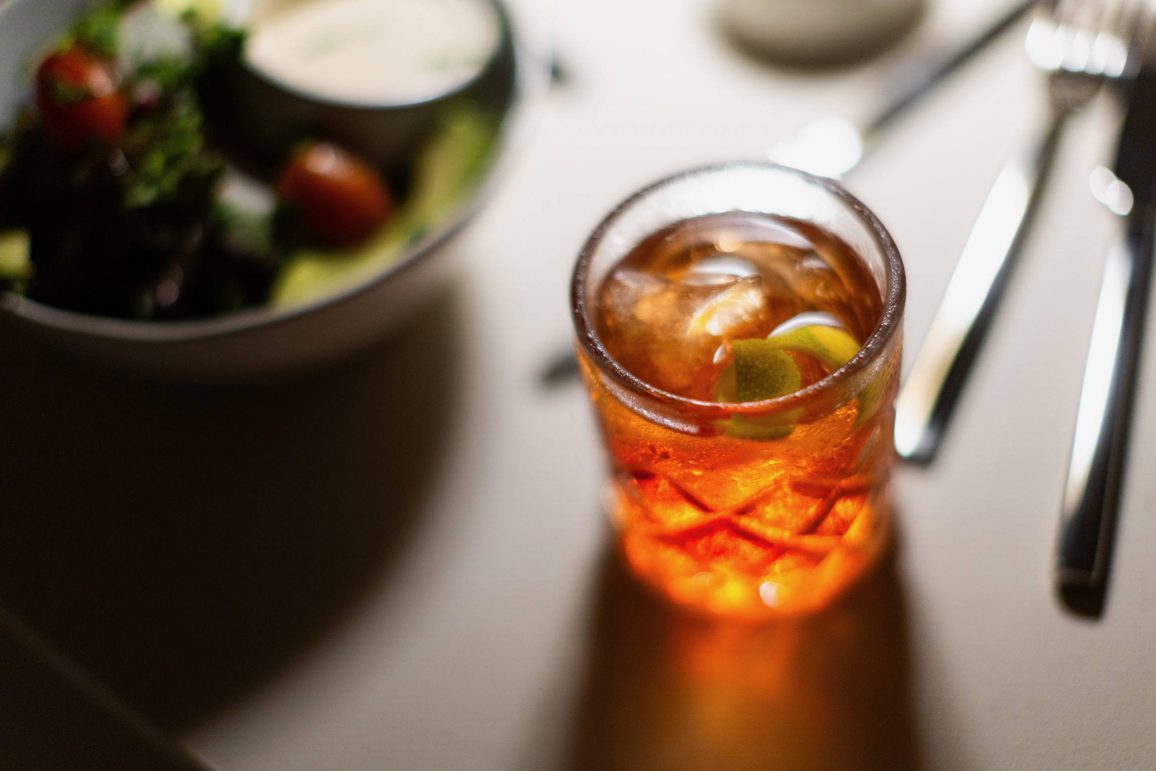 A glass of iced tea with lemon slices on a table next to a bowl of salad with cherry tomatoes and leafy greens.