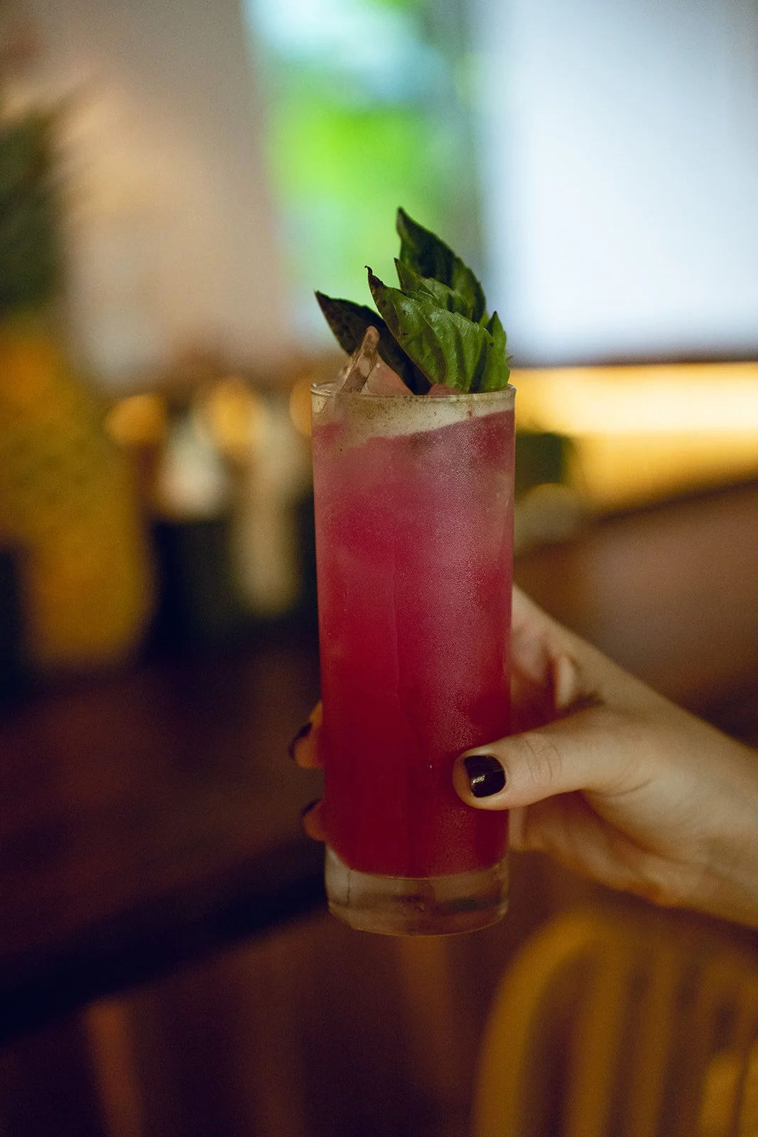 A hand with black painted nails holding a tall glass of pink cocktail garnished with fresh basil leaves, indoors with blurred warm lighting in the background.