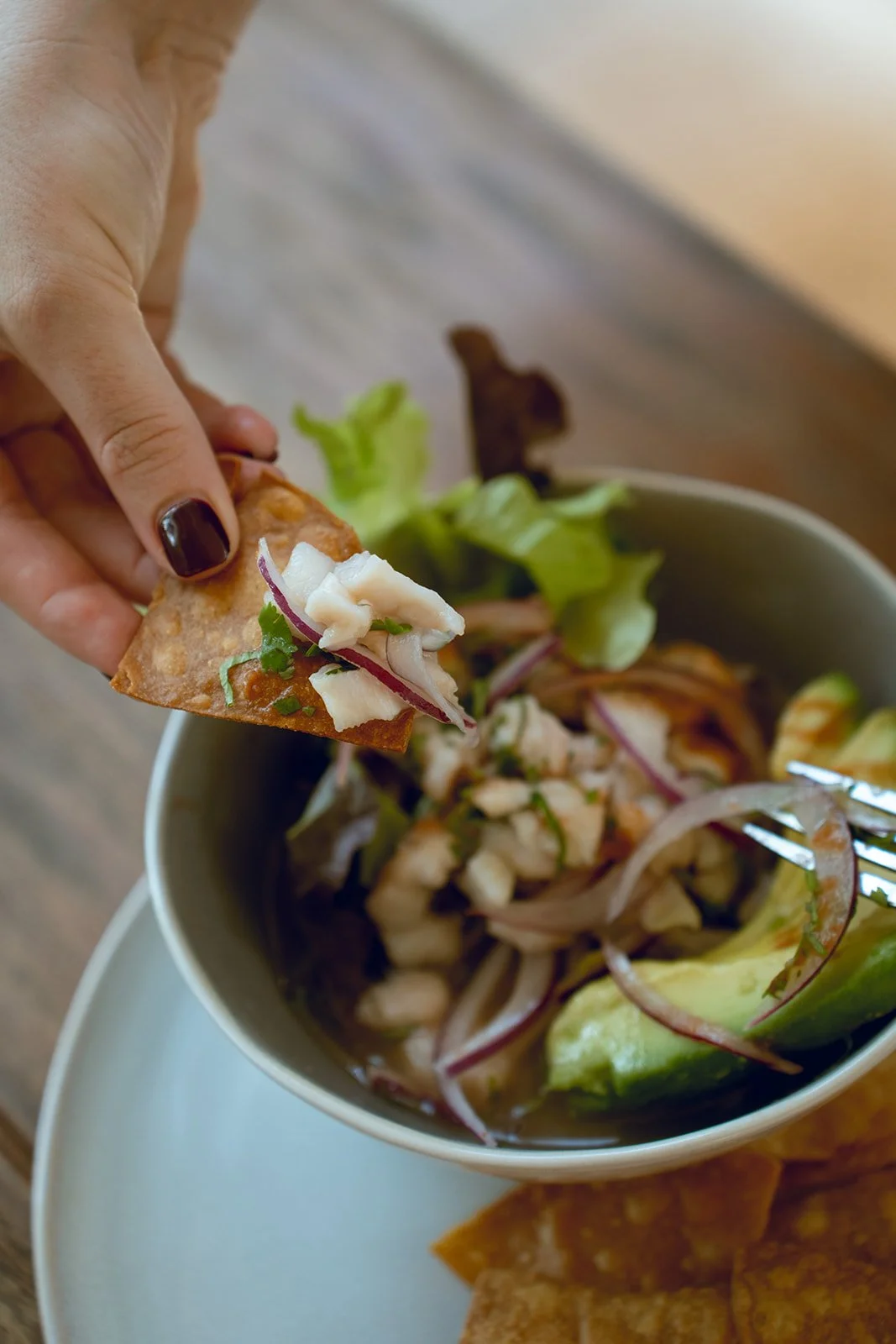 Person holding a tortilla chip with salsa and seafood, dipping into a bowl of seafood salad with lettuce and onions, served with tortilla chips on the side.