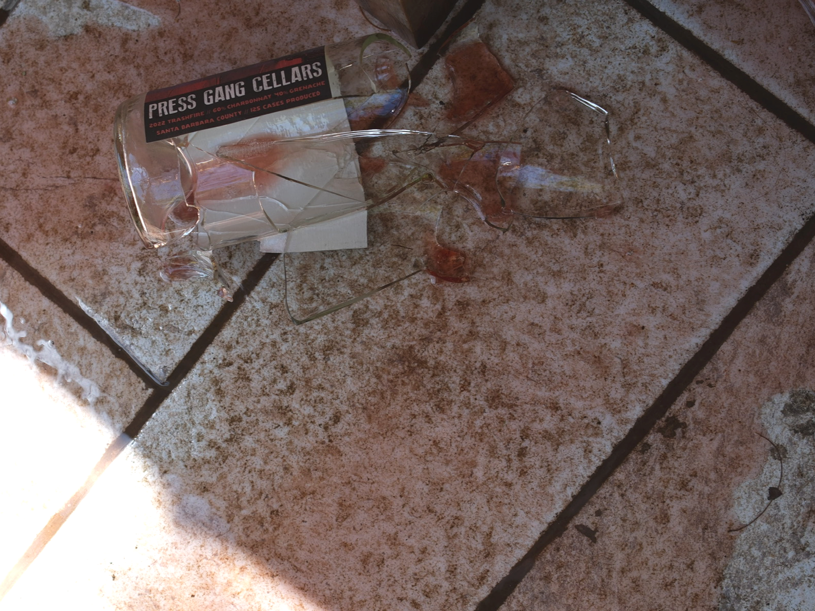Broken glass upside down on a tiled floor with a bottle labeled 'Press Gang Cellars' nearby.