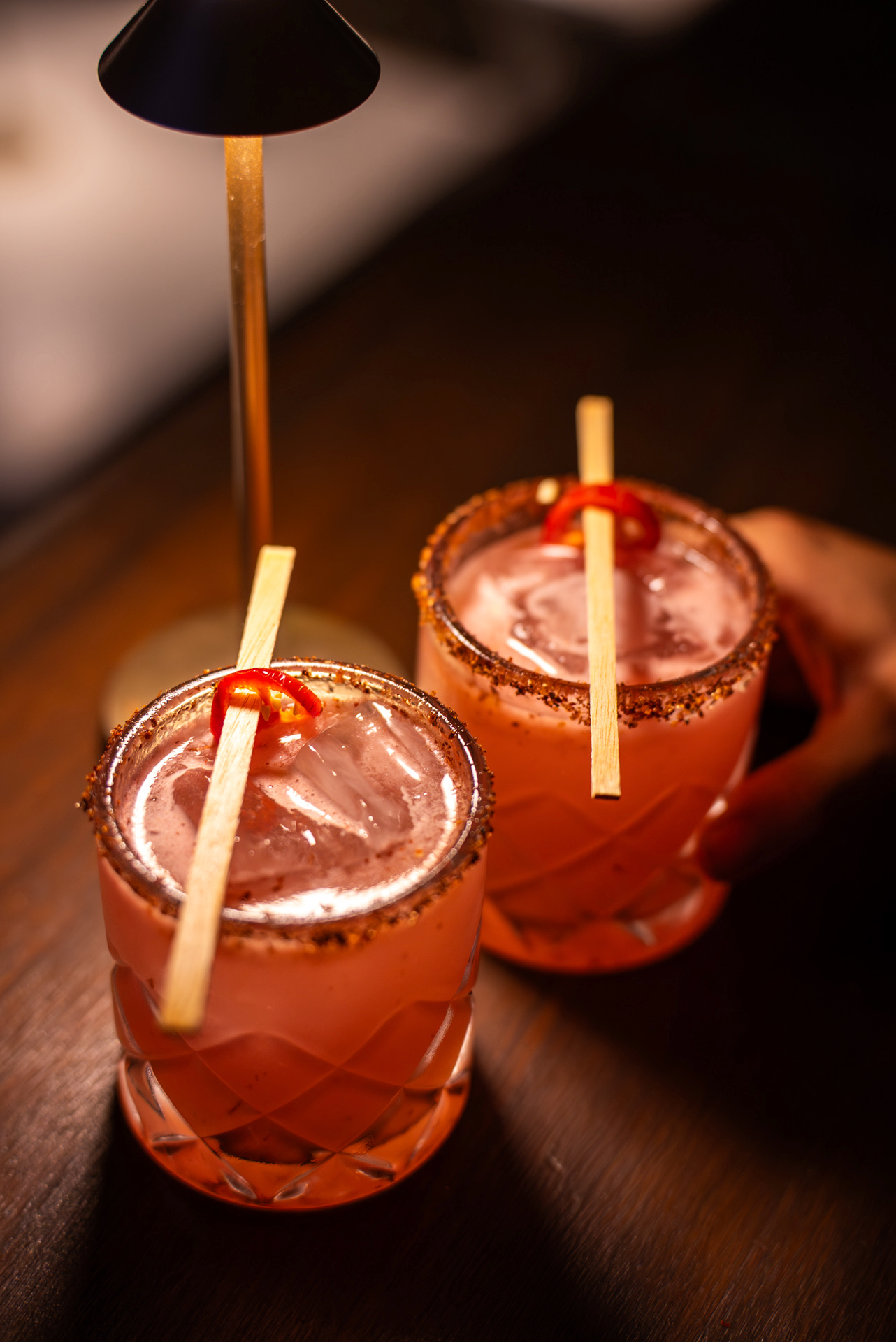 Three pink cocktails with salted rim, garnished with red chili peppers and served with wooden sticks, on a wooden surface