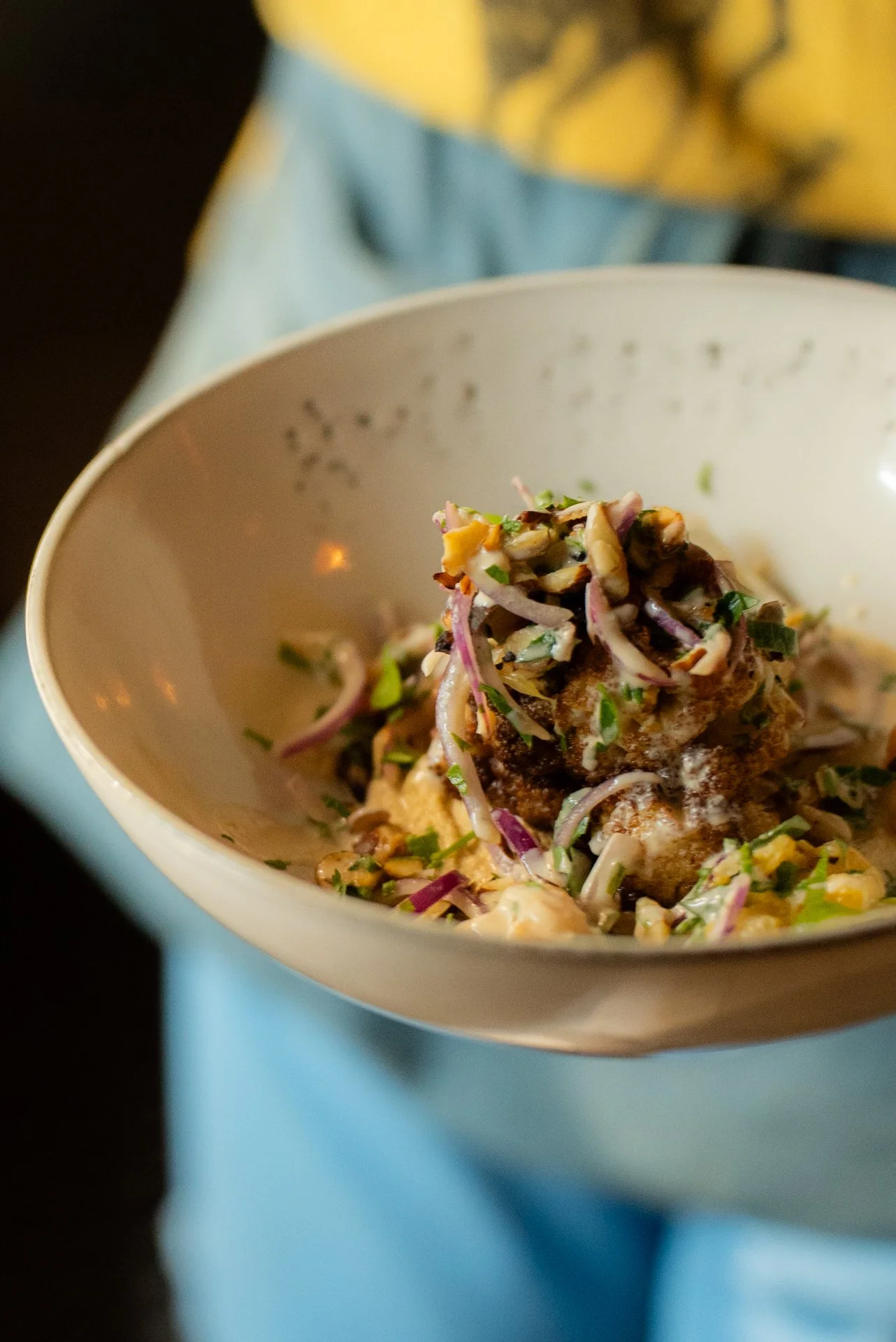 Close-up of a bowl containing a food dish topped with chopped herbs, vegetables, and a seasoned meatball, with a plate of potatoes in the background.