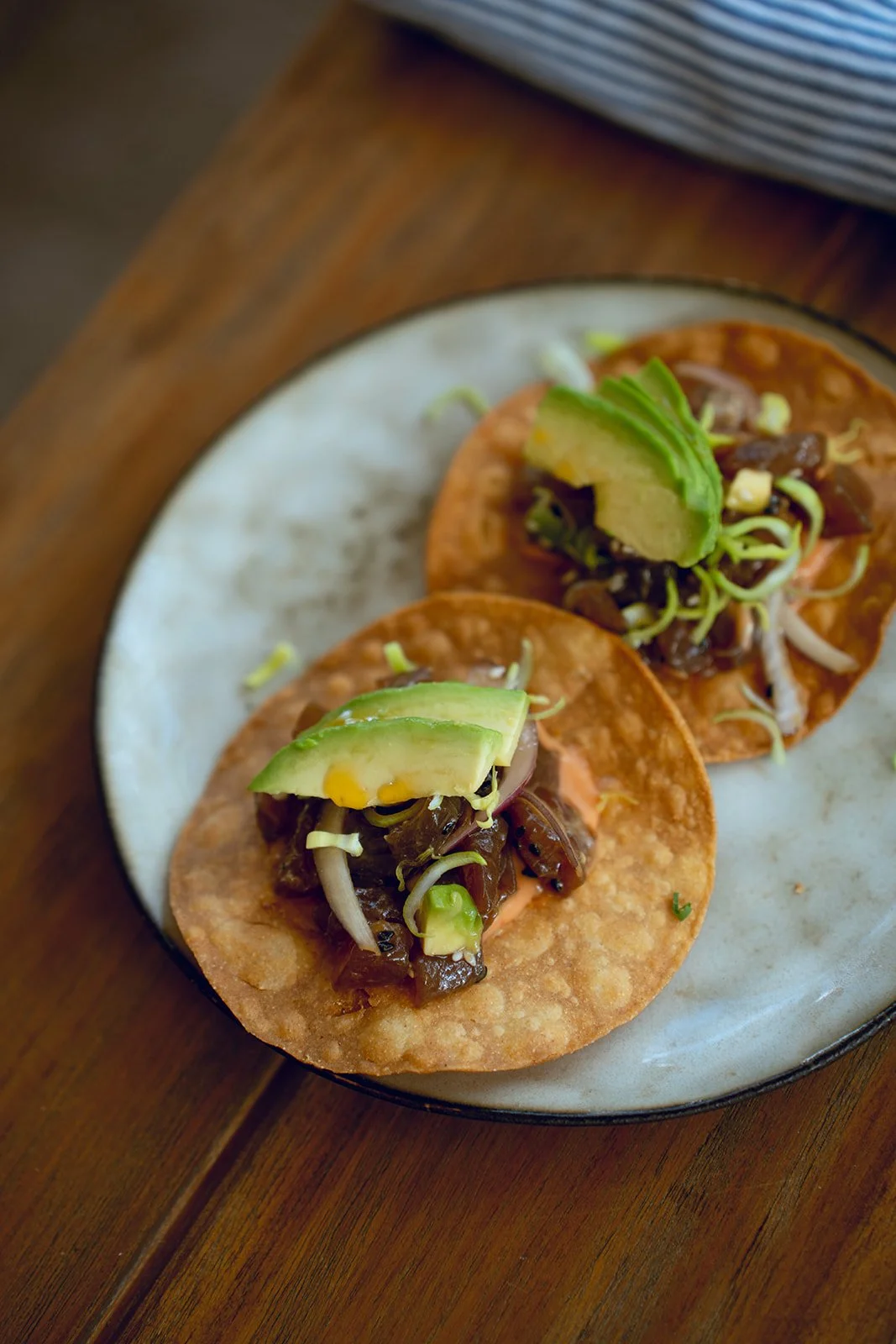 Close-up of two street-style tacos on a white plate, topped with avocado slices and garnished with shredded lettuce and sauce, resting on a wooden table.