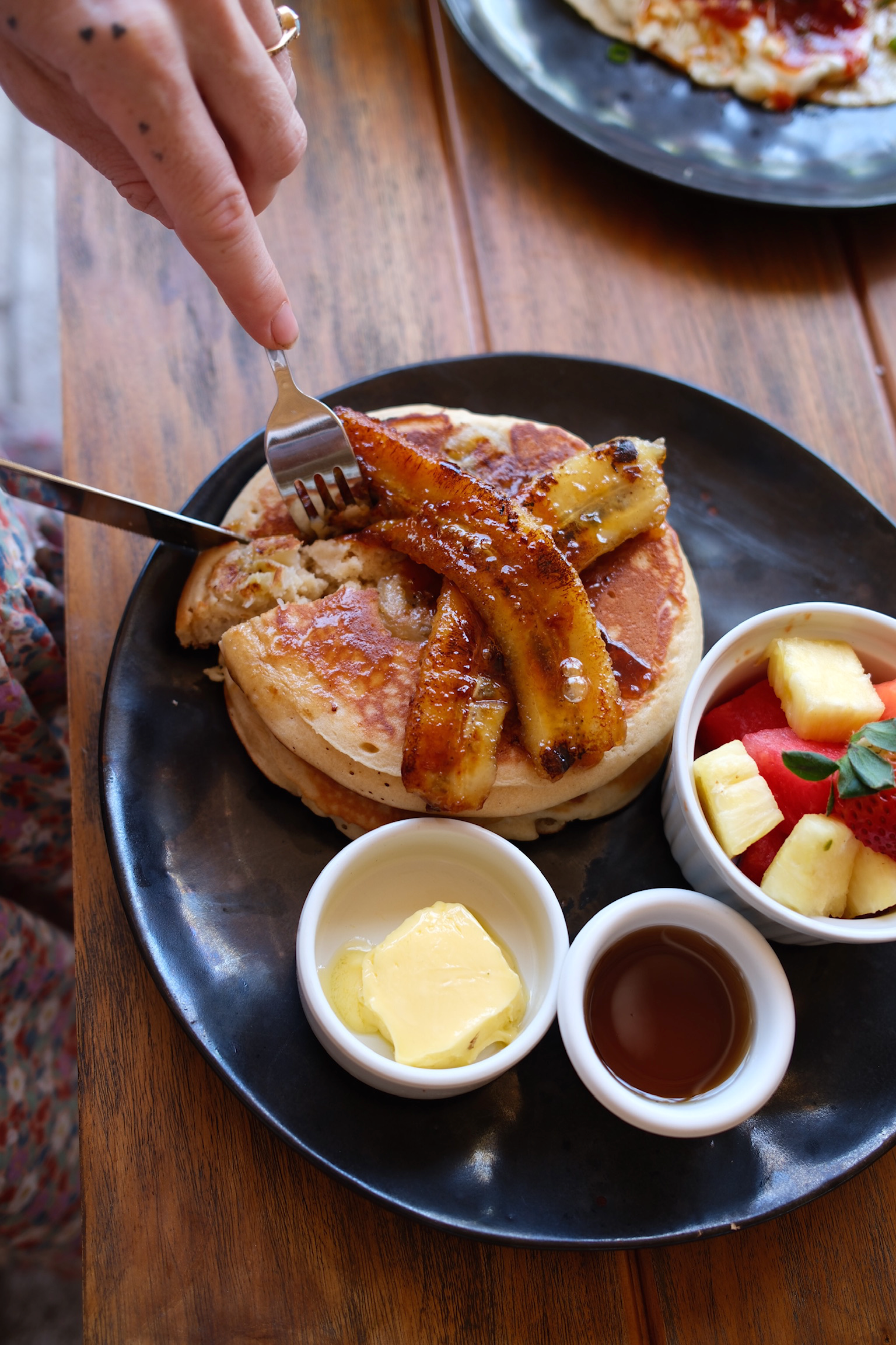 Close-up of a breakfast dish with a biscuit topped with caramelized bananas and syrup, served on a black plate with side bowls of butter, syrup, and fresh fruit salad on a wooden table.