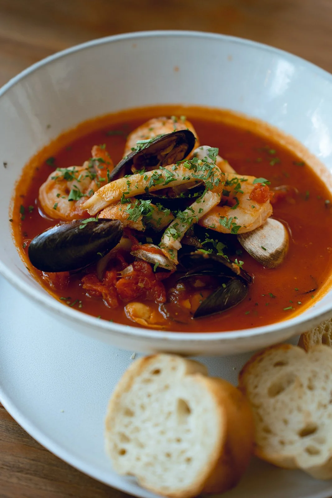 A bowl of seafood soup with mussels, shrimp, and fish, garnished with herbs, served with slices of bread.