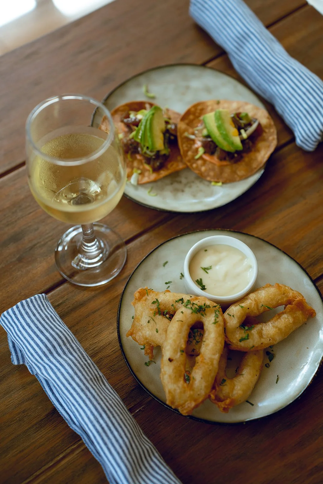 A glass of white wine, a plate with two tacos topped with avocado, and a plate of fried calamari with a dipping sauce on a wooden table with a blue and white striped napkin.