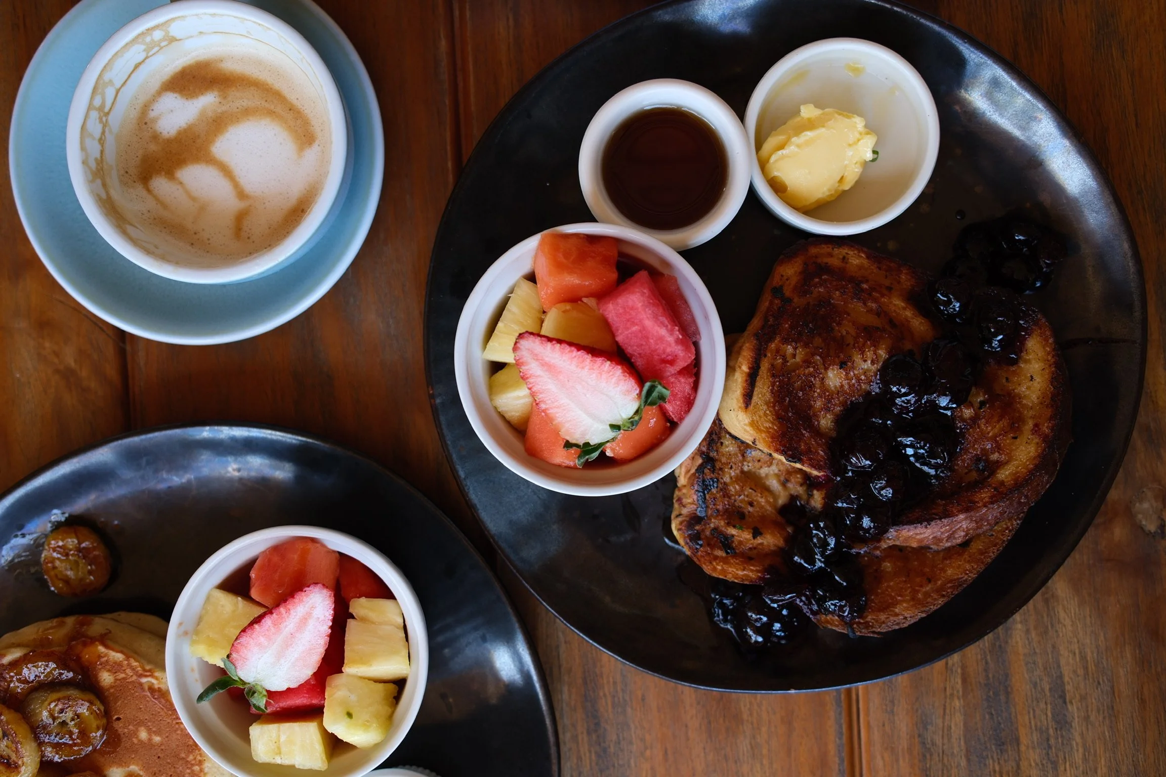 A breakfast plate with French toast topped with blueberries, served with side dishes of butter, syrup, and fresh fruit, accompanied by a cup of coffee.