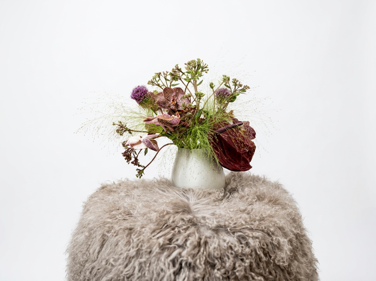 Flower arrangement with pink, purple, and leafy green flowers in a white vase on a furry beige surface against a plain white background.