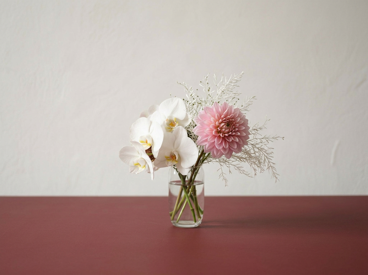 A clear glass vase containing white orchids, a pink dahlia, and some dried white and pale green foliage, placed on a maroon surface against a plain light-colored wall.