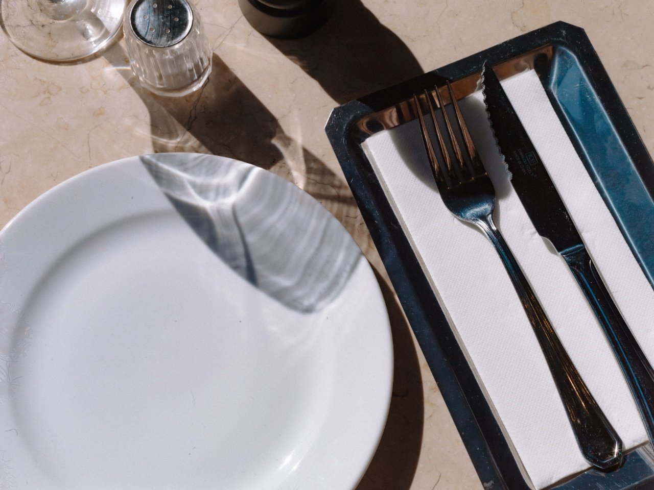 Empty white plate and utensils on a paper napkin inside a black tray, with salt and pepper shakers and a small container nearby on a beige marble table.